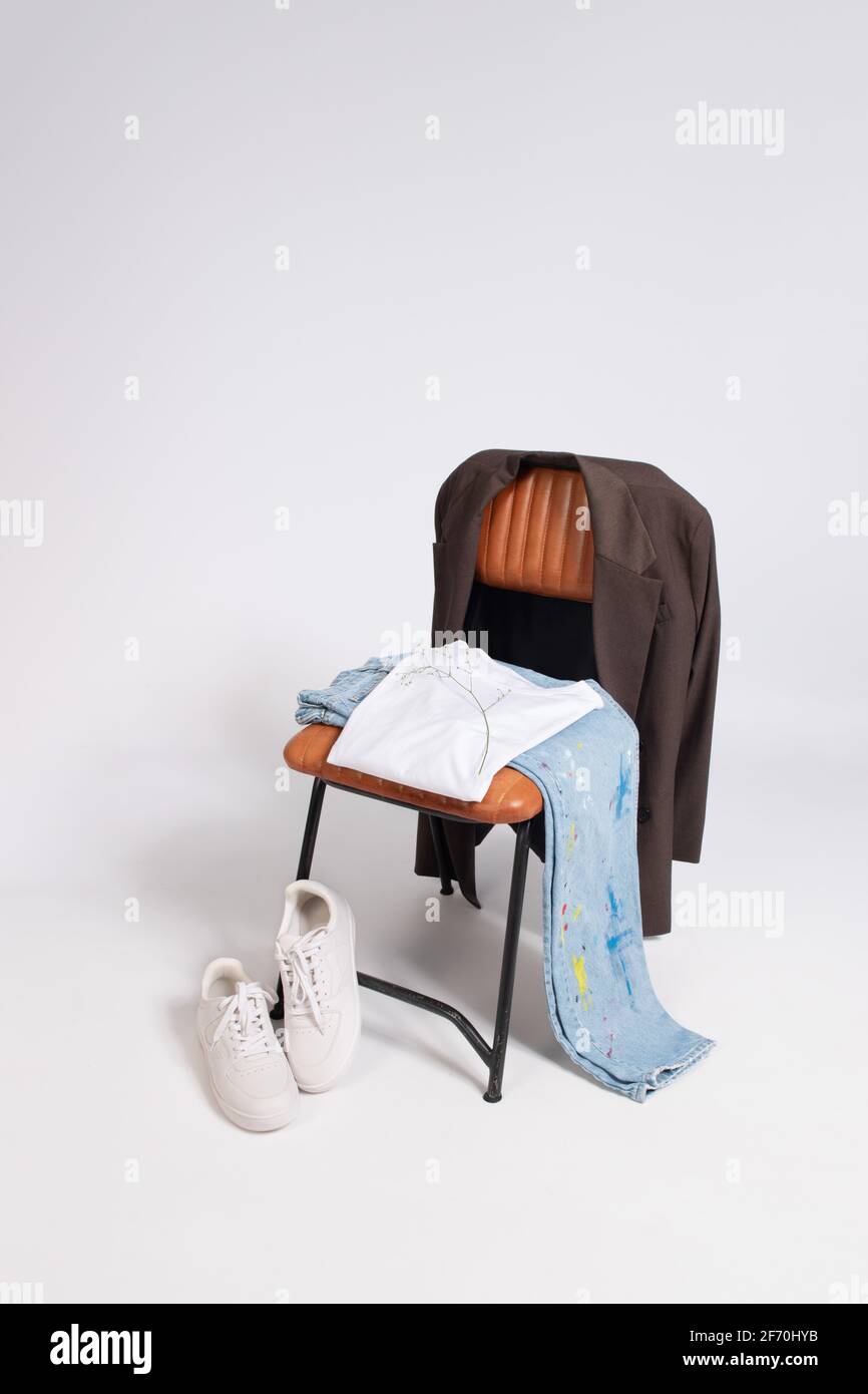 Sneakers bianche, jeans blu, t-shirt bianca posata su una sedia in pelle marrone e metallo nero con una giacca marrone scuro adagiato sul retro della sedia. wh Foto Stock