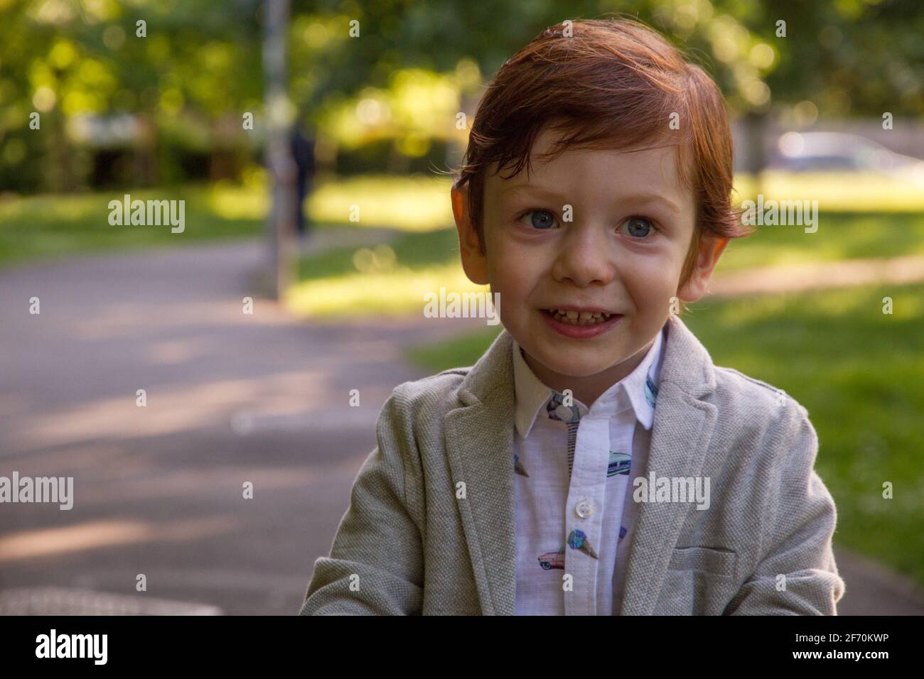 Ritratto di un simpatico ragazzo rosso dagli occhi blu che indossa un blazer grigio e una camicia bianca in un parco in una giornata di sole Foto Stock