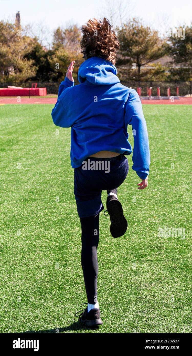 Una vista posteriore di un runner della scuola superiore che pratica la velocità e l'agilità pratica gli esercizi sportivi su un campo di erba verde con i suoi capelli che rimbalzano su e giù. Foto Stock