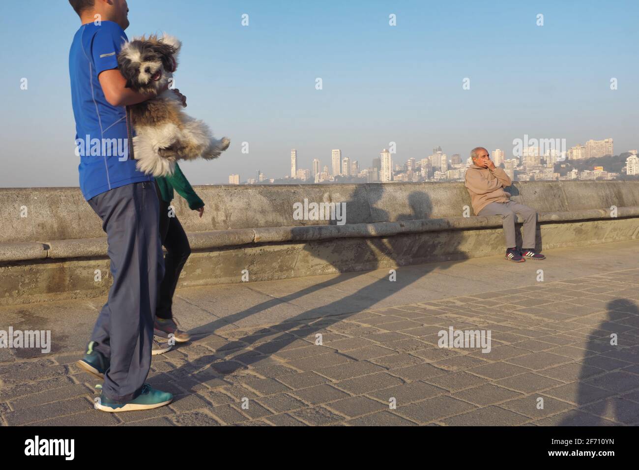 Un uomo porta il suo cane da compagnia lungo Marine Drive, un viale sul mare vicino al Mar Arabico a Mumbai, India, un uomo seduto in relax sullo sfondo Foto Stock