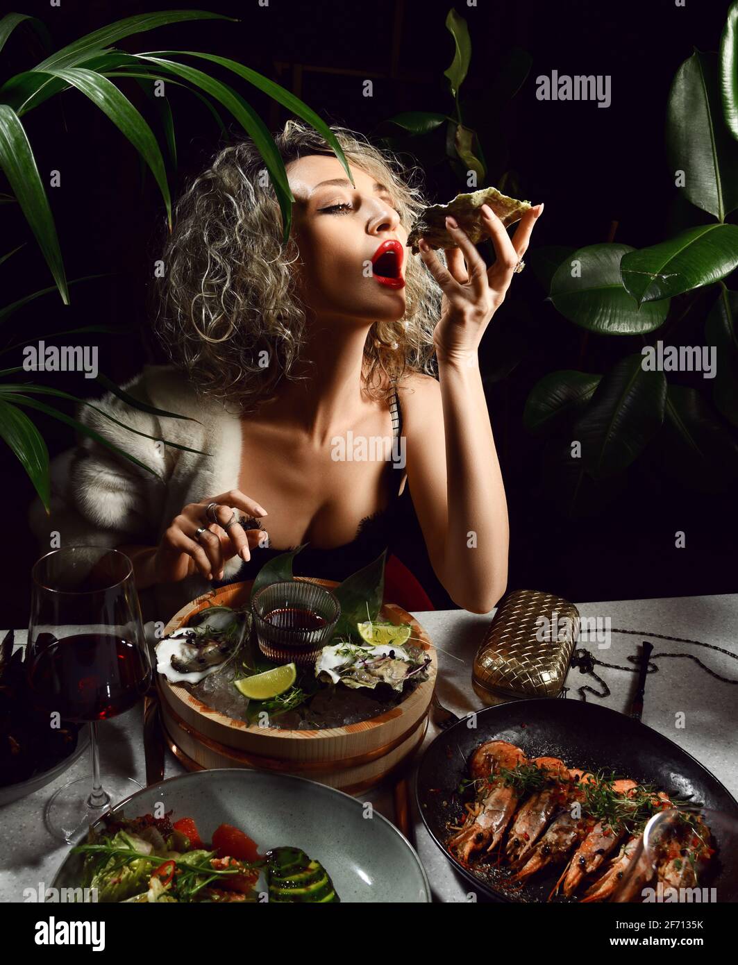 Giovane donna bionda in abito da sera con scollo profondo mangia ostriche di pesce, gamberi, cozze e insalata nel ristorante Foto Stock