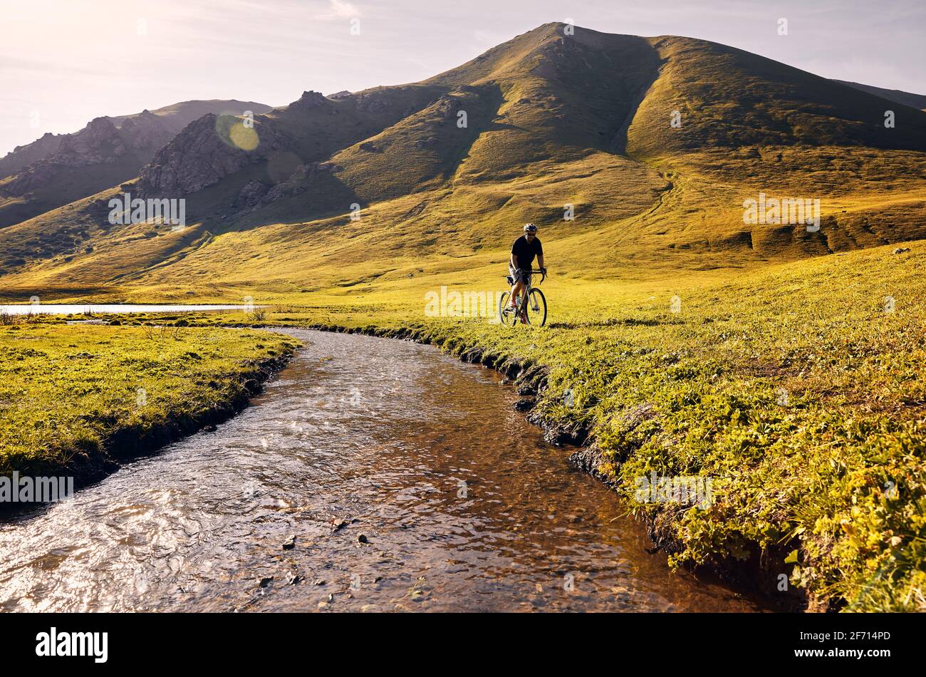 L'uomo in mountain bike corre vicino al fiume nella verde valle di montagna all'alba. Concetto di ricreazione, viaggio e stile di vita sano. Foto Stock