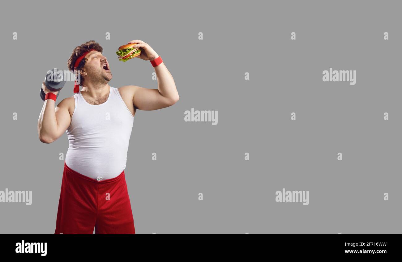 Divertente sportivo caucasico in sovrappeso tenendo dumbbell mangiare hamburger studio shot Foto Stock