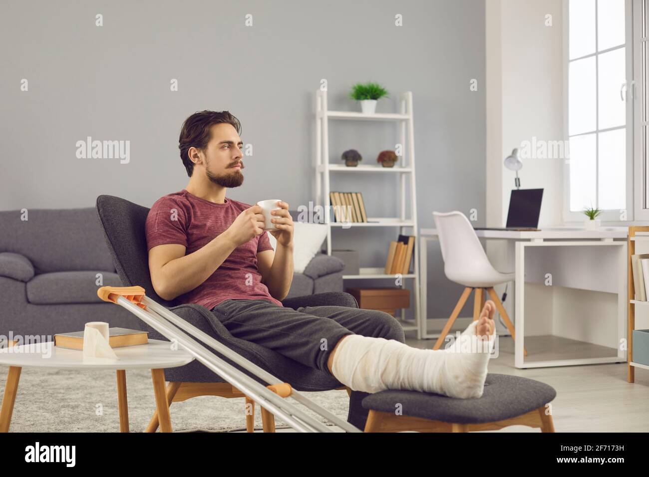 Giovane uomo con frattura dell'osso della gamba che si sente triste e annoiato mentre rimaniate da soli a casa Foto Stock