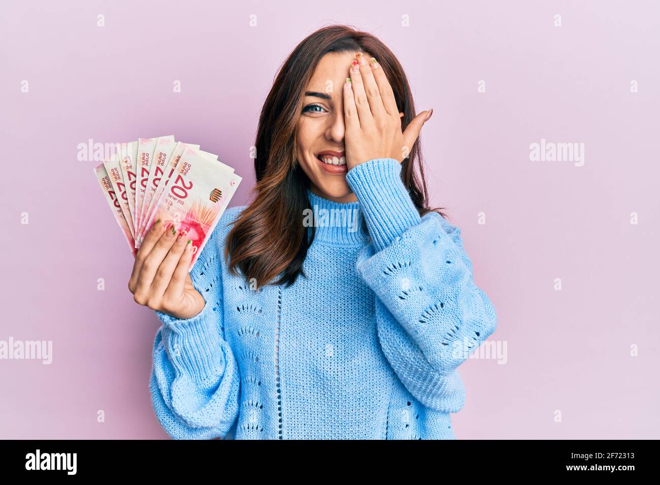 Giovane donna di brunetta che tiene 20 israel shekels banconote che coprono un occhio con la mano, sorriso fiducioso sul viso e sorpresa emozione. Foto Stock