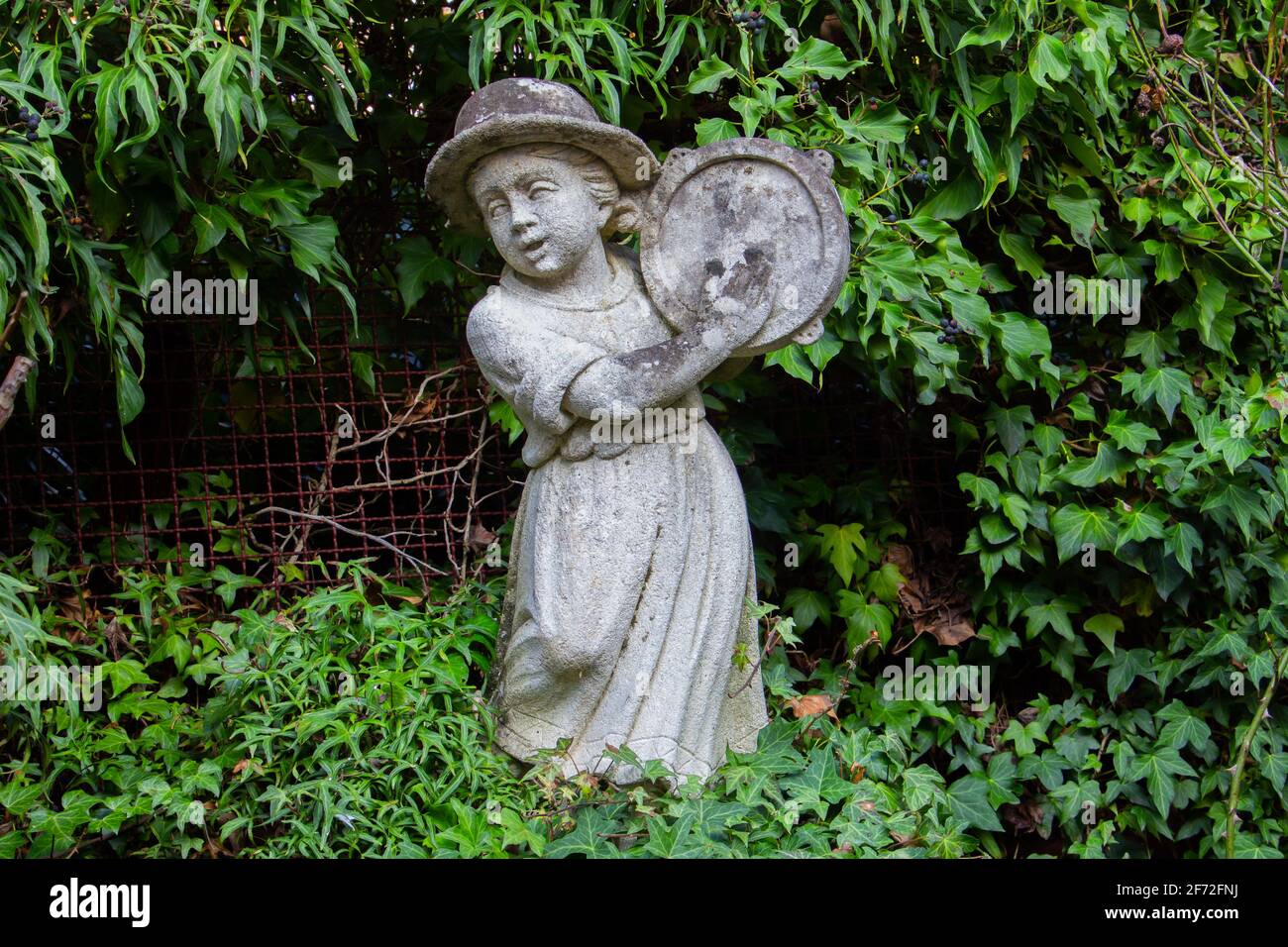 Vecchia statua bianca intemperie di una ragazza che gioca un tamburello circondato da edera Foto Stock
