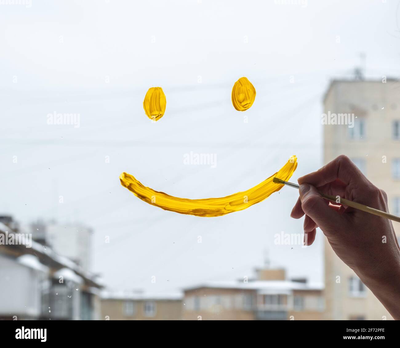 Una mano di donna disegna un volto sorridente con vernice gialla sulla finestra di vetro con una vista delle case della città, primo piano. Un simbolo di speranza, felicità, gioia. Foto Stock