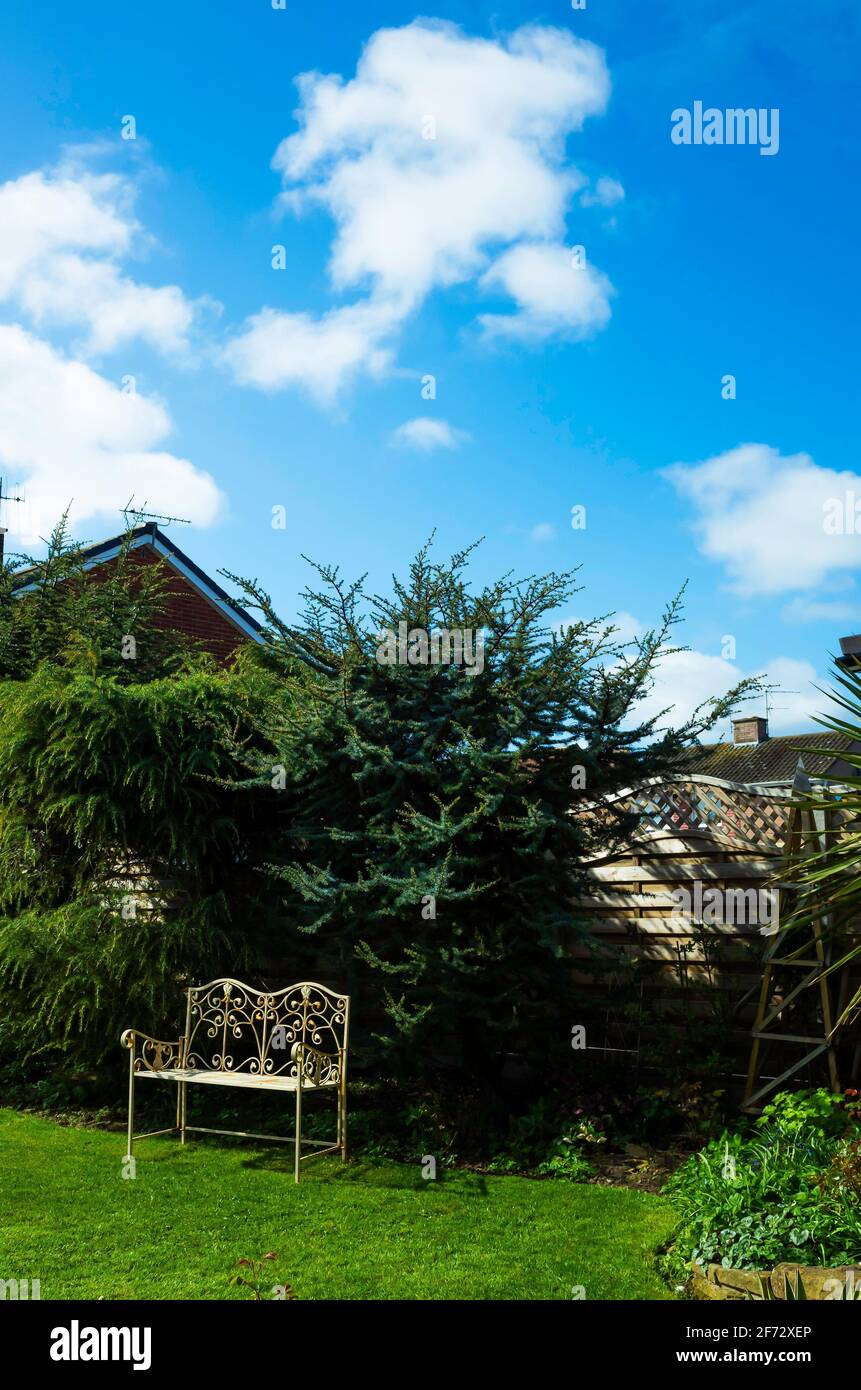 Una panca da giardino in ferro battuto dipinta di giallo in un angolo soleggiato di un giardino domestico con cielo blu e. nuvole bianche Foto Stock