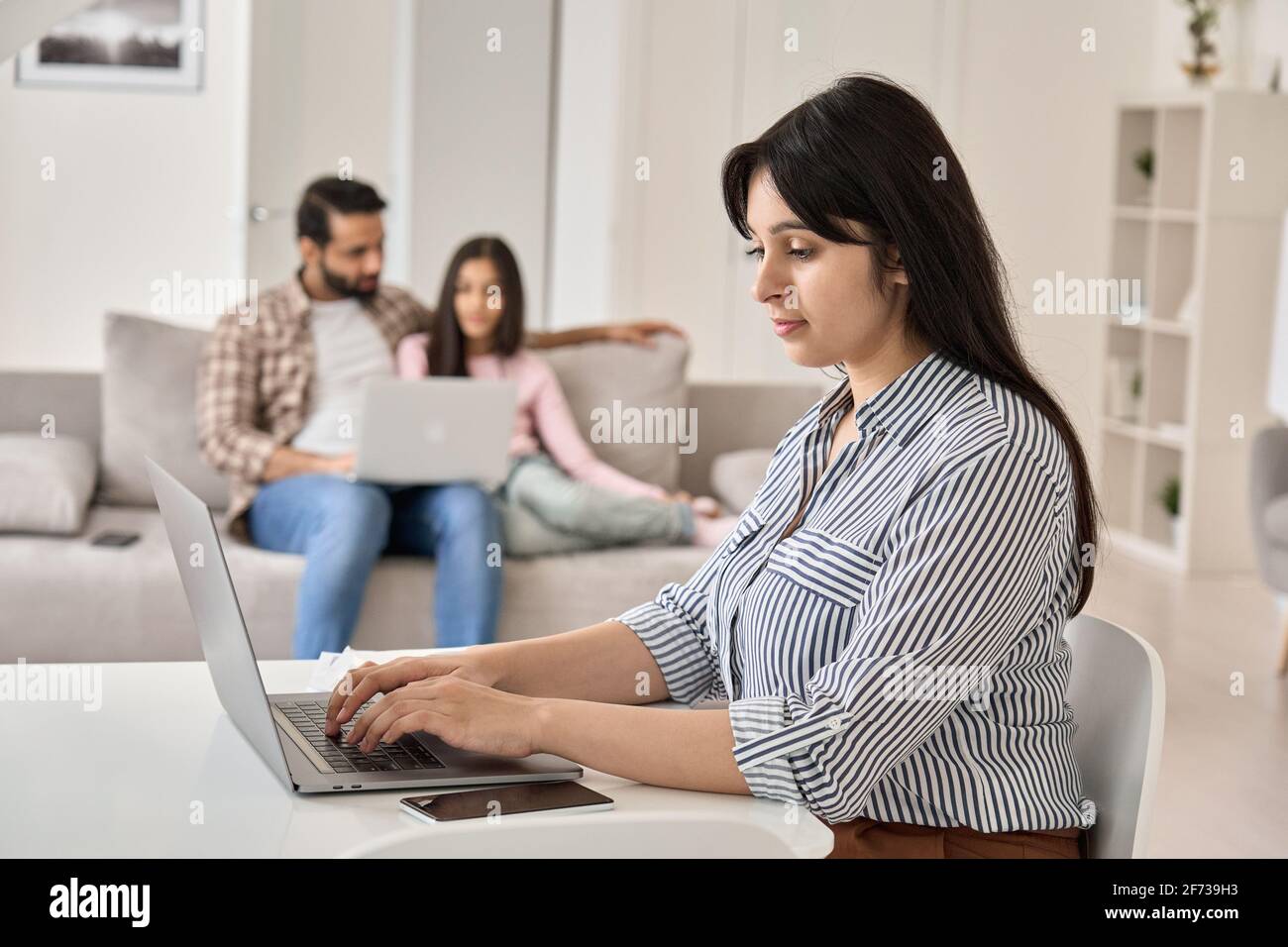 Giovane donna indiana che lavora a distanza in linea dall'ufficio domestico con la famiglia. Foto Stock