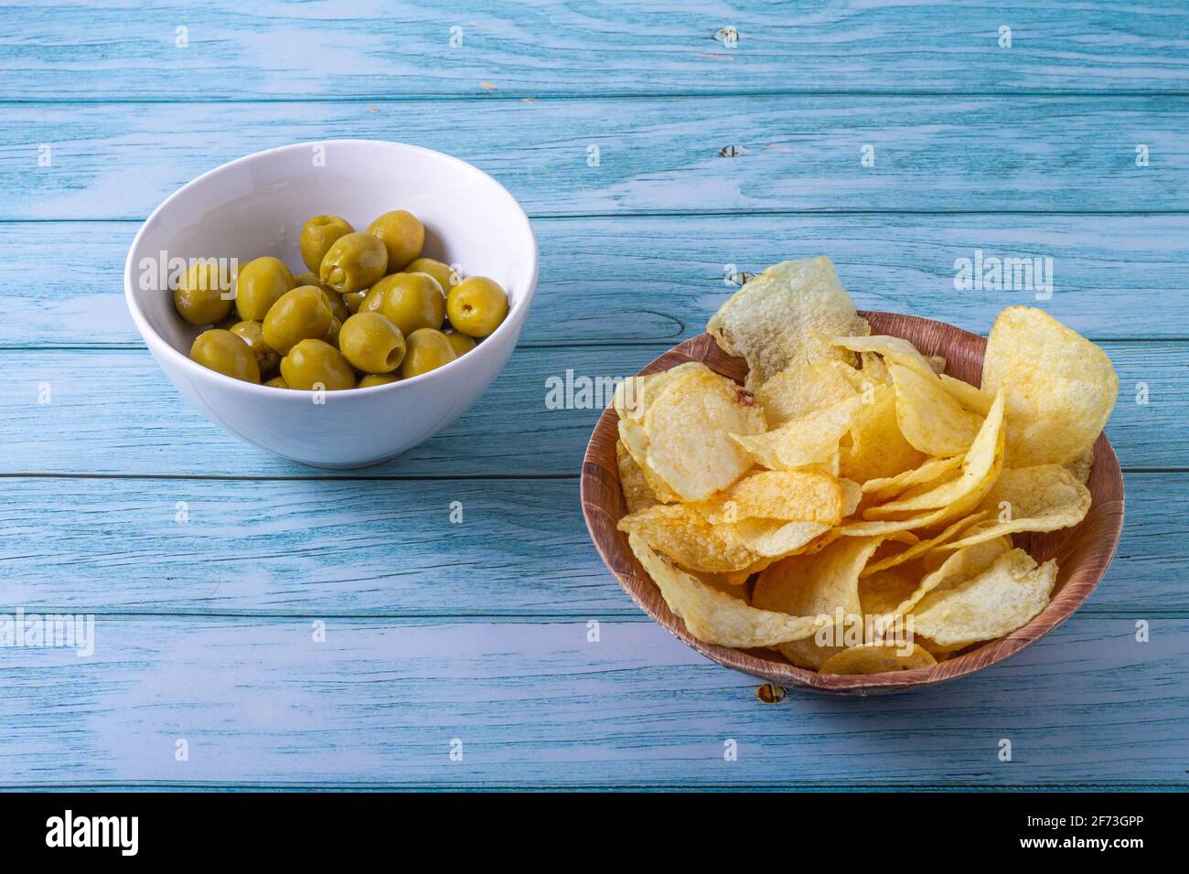 Concetto di spuntino spagnolo. Olive ripiene in una ciotola bianca e patatine in una ciotola di legno su un tavolo di legno in tonalità blu Foto Stock