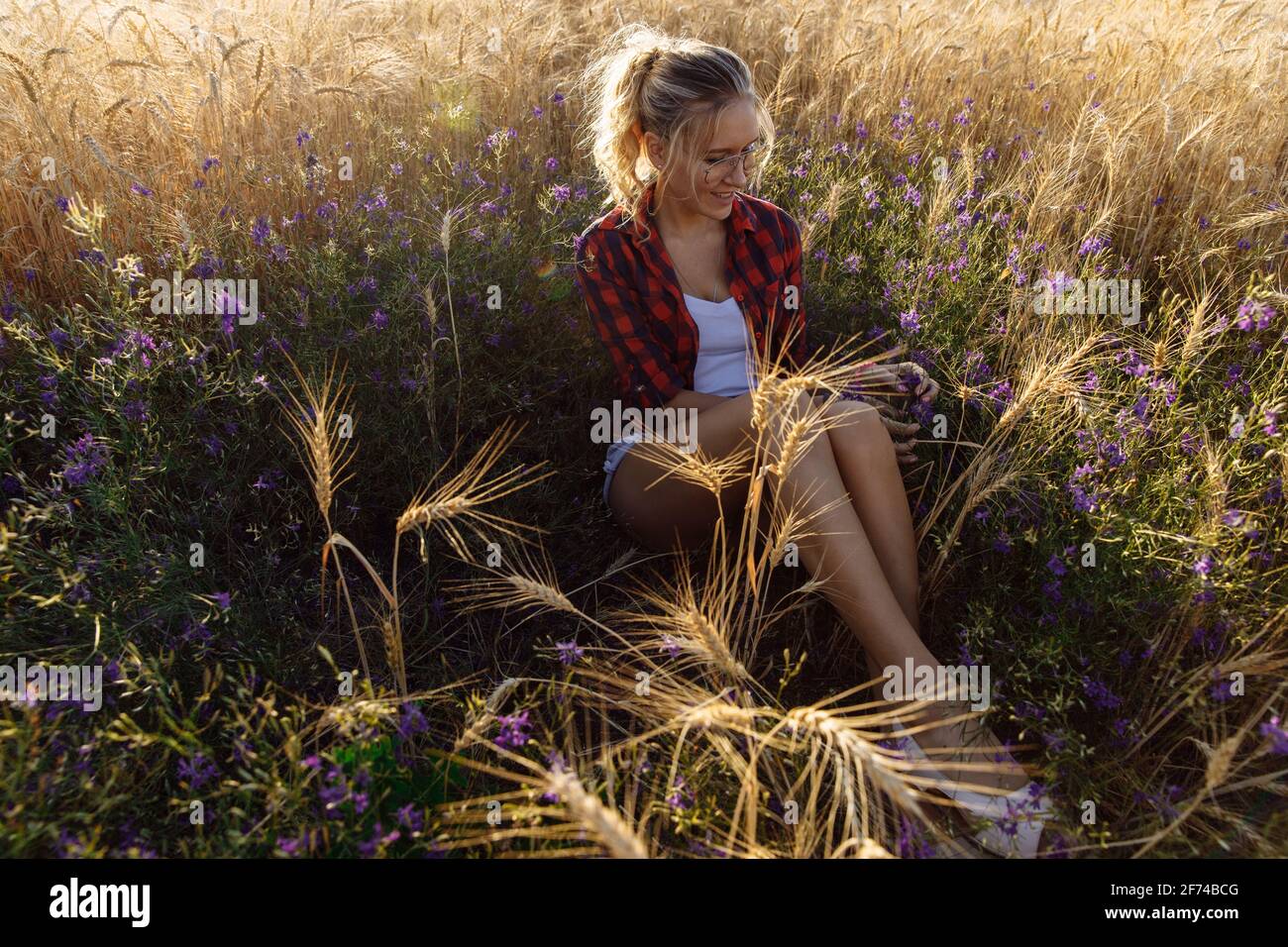 Una giovane donna al tramonto si siede in un campo con fiori e grano. Foto Stock
