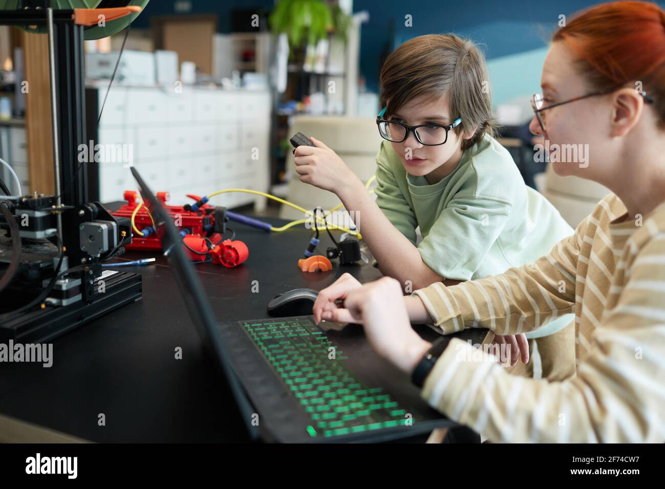 Vista laterale di una giovane insegnante che aiuta il ragazzo utilizzando il 3D stampante durante la lezione di robotica e ingegneria a scuola Foto Stock