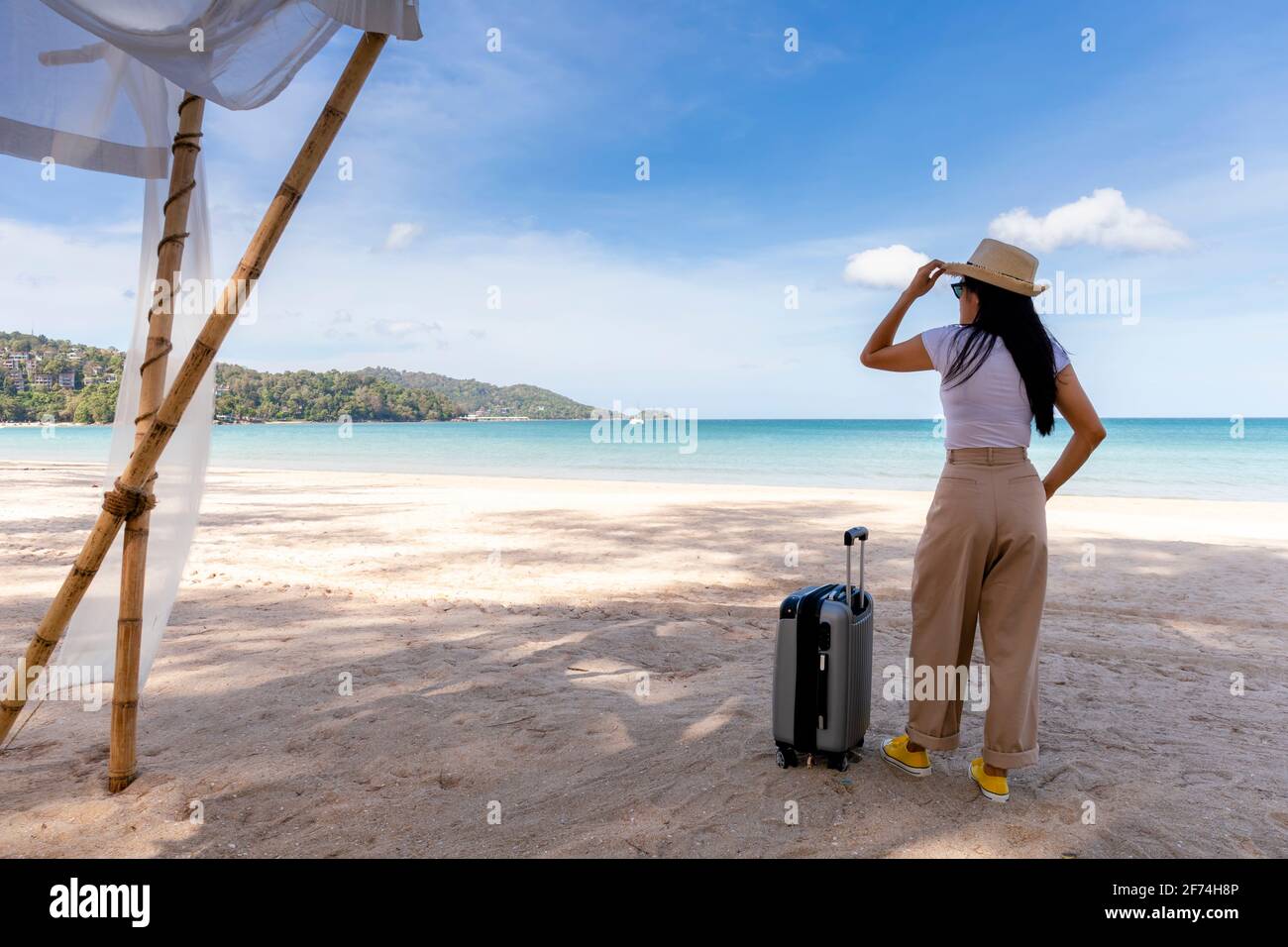 Bella giovane donna indossare T-shirt, pantaloni lunghi e cappello di  paglia con una valigia su una spiaggia tropicale. Mare blu sullo sfondo.  Felice viaggio donne stand Foto stock - Alamy