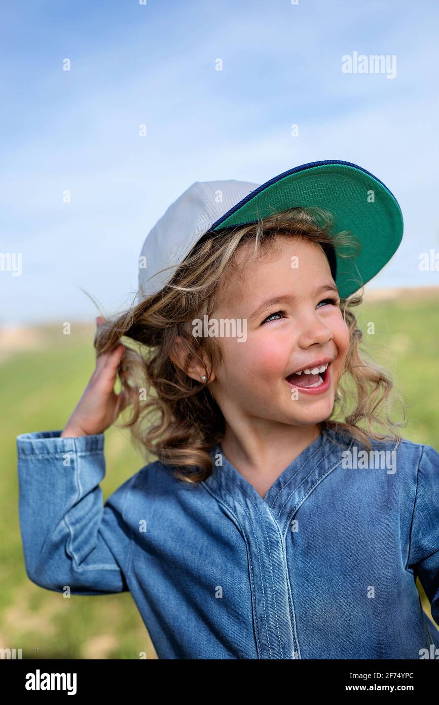 Ragazza bionda felice in cappello che tocca la testa e che guarda via contro il cielo blu in estate in prato Foto Stock