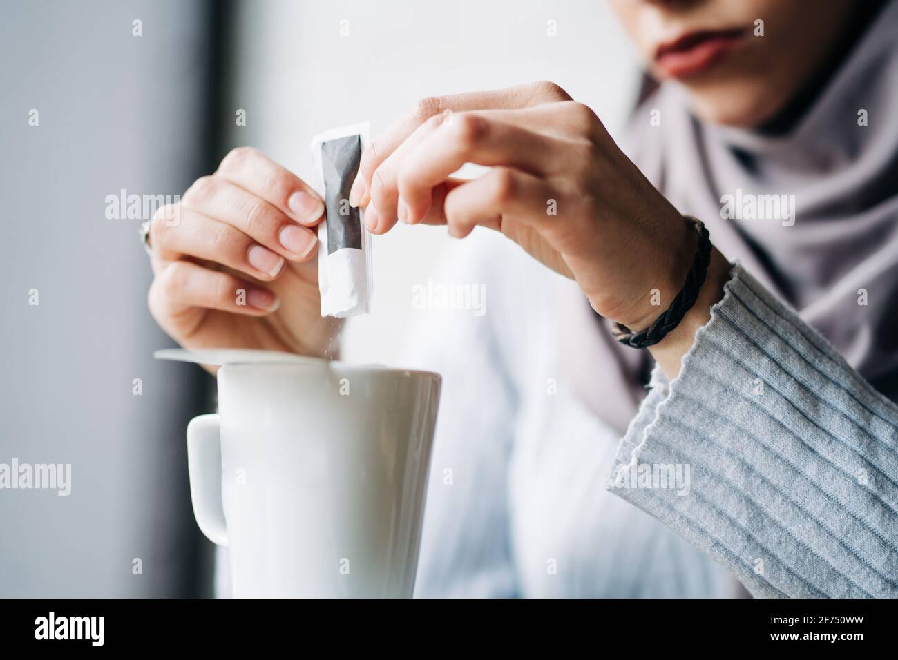 Crop anonimo musulmano femmina in hijab versando zucchero da carta pacchetto in tazza con bevanda calda al caffè Foto Stock