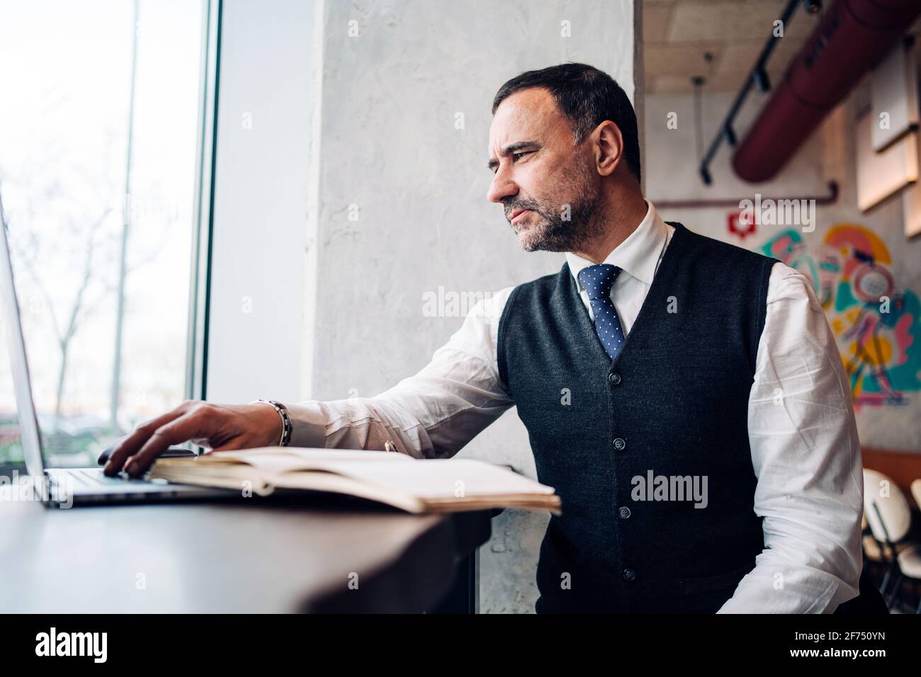 Imprenditore maschio ispanico maturo in abiti formali che lavorano su netbook al tavolo da caffè con diario Foto Stock