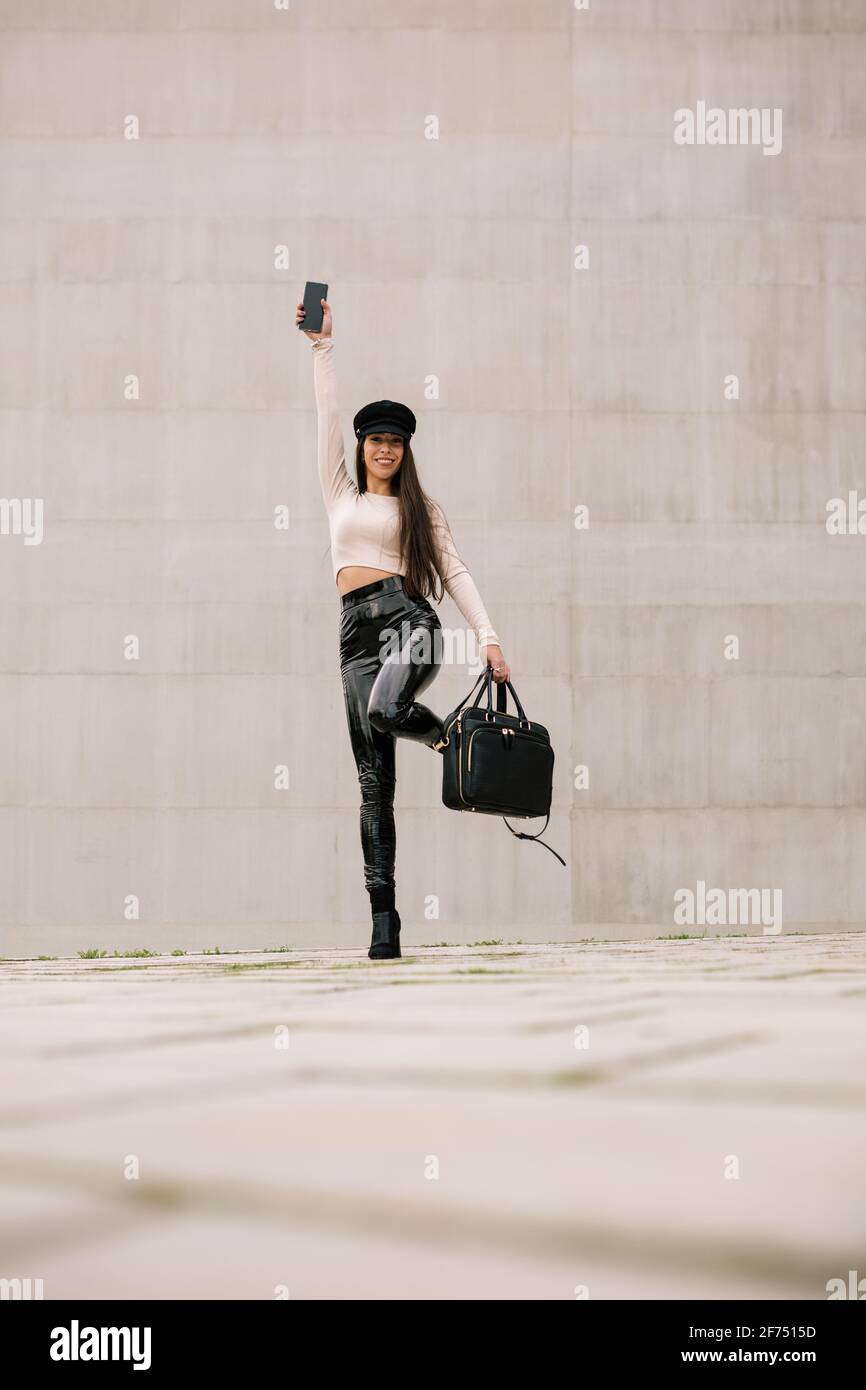 Piano terra di imprenditrice femminile deliziato in abiti alla moda e. con smartphone in piedi sulla strada della città e guardando la fotocamera Foto Stock