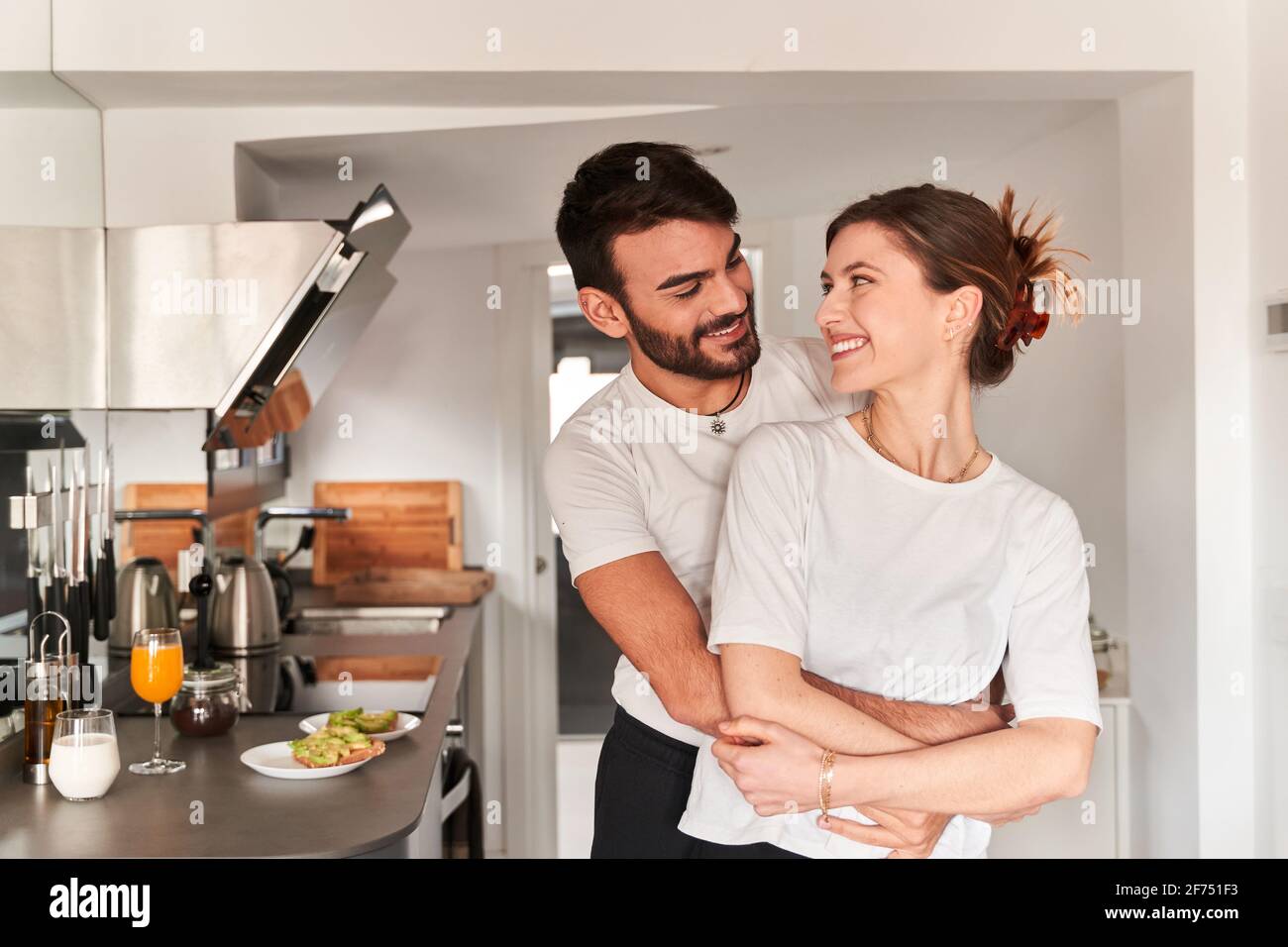 Vista laterale di un giovane ragazzo etnico romantico in bianco t camicia sorridente e abbracciante ragazza felice mentre si guarda ciascuno altro Foto Stock