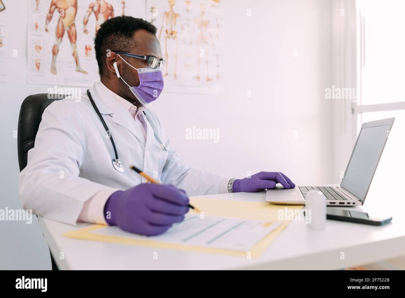 Medico afroamericano in occhiali che lavora con il paziente in linea sopra netbook durante la scrittura del file paziente sul tavolo dell'ospedale Foto Stock