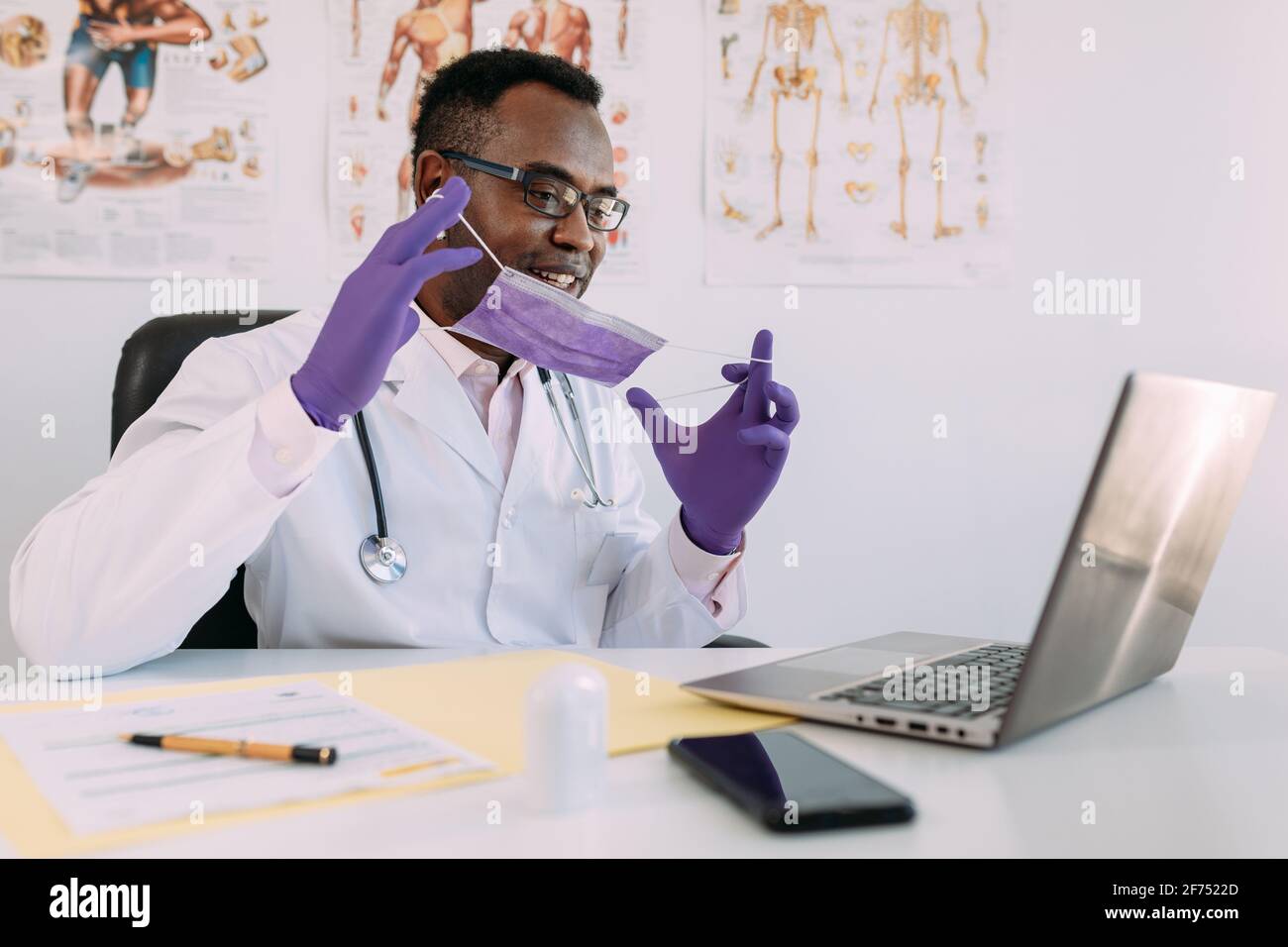 Medico afroamericano in occhiali e maschera facciale che lavora con paziente online su netbook durante la scrittura del file paziente all'indirizzo tavolo in ospedale Foto Stock