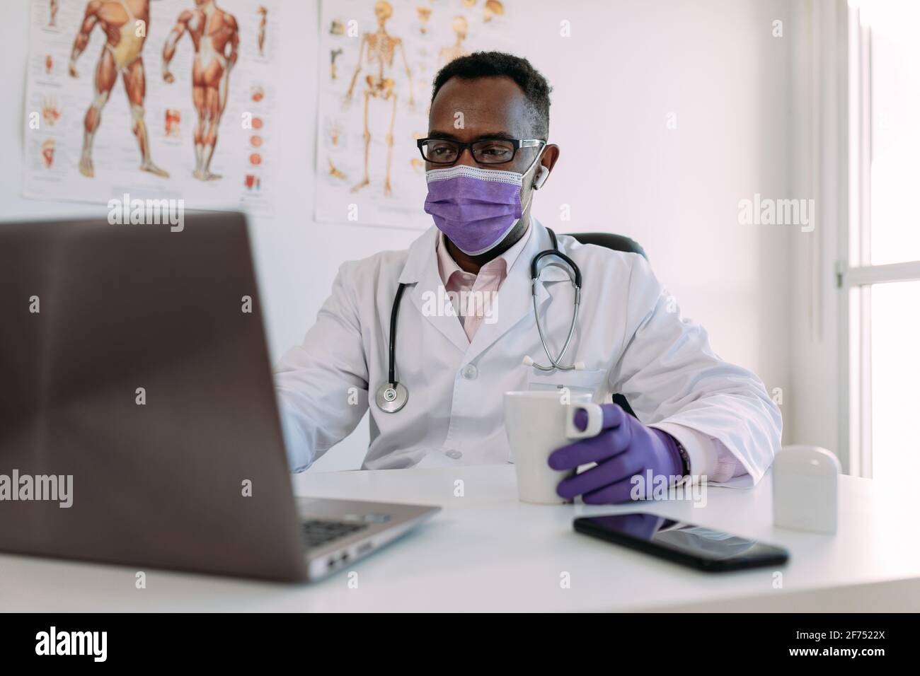 Irriconoscibile concentrato africano americano medico maschile in accappatoio medico e. maschera bere caffè e lavorare su computer portatile in clinica moderna Foto Stock