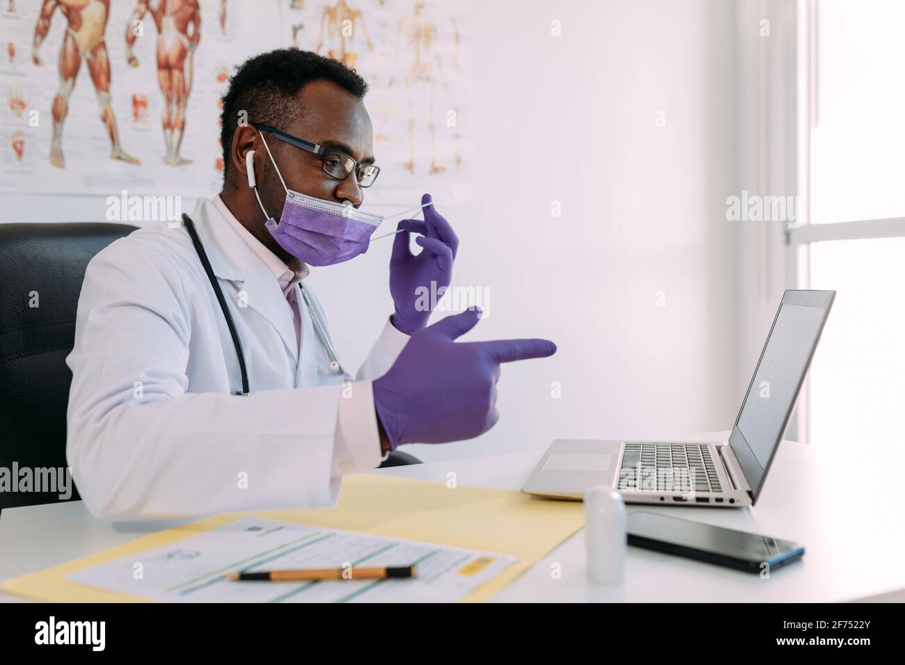 Medico afroamericano in occhiali e maschera facciale che lavora con paziente online su netbook durante la scrittura del file paziente all'indirizzo tavolo in ospedale Foto Stock
