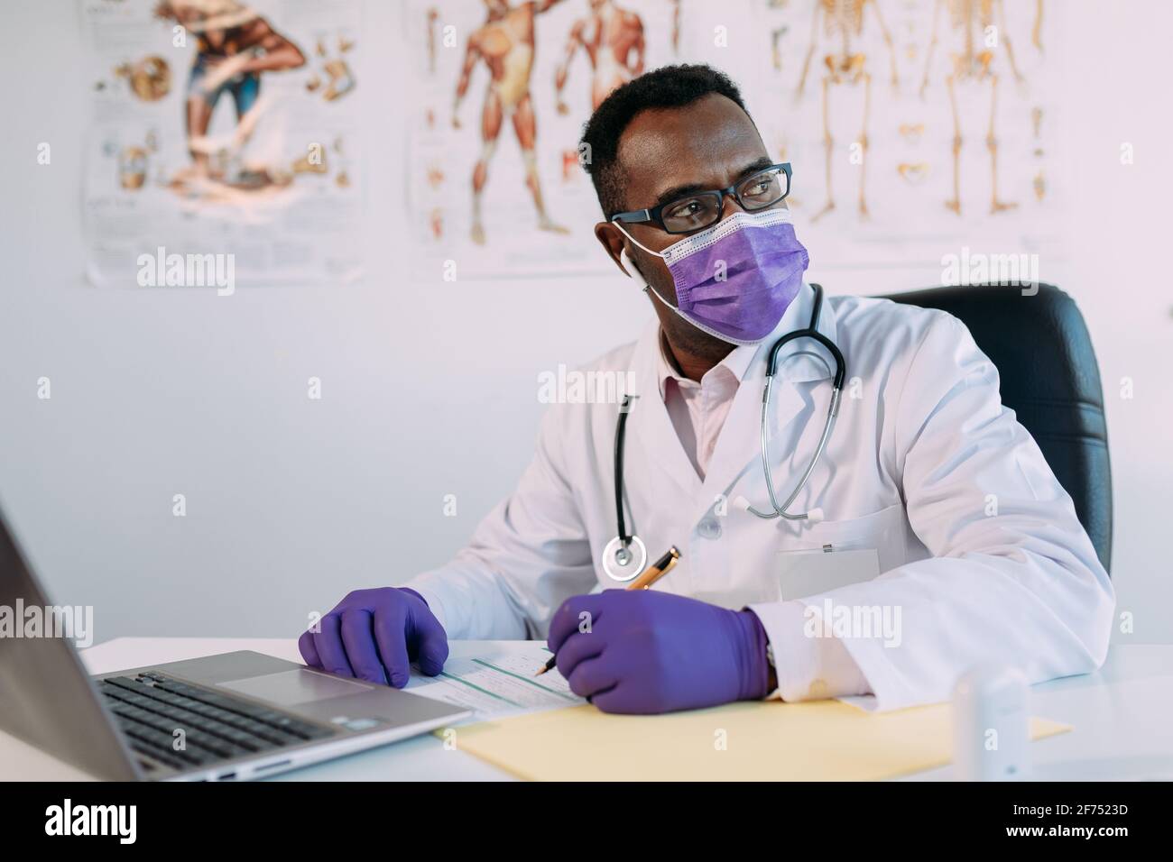 Medico afroamericano in occhiali che lavora con il paziente in linea sopra netbook durante la scrittura del file paziente sul tavolo dell'ospedale Foto Stock