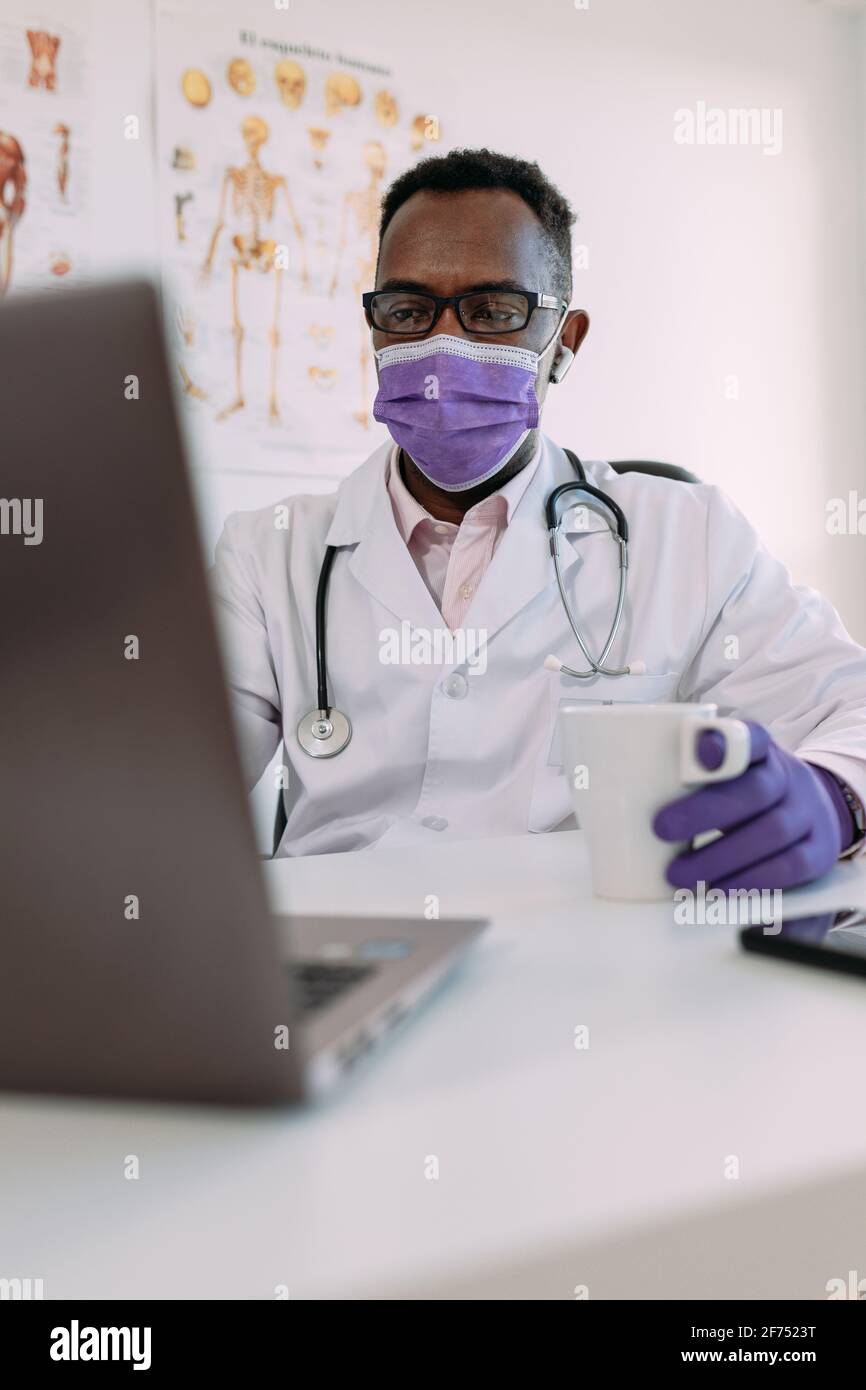 Irriconoscibile concentrato africano americano medico maschile in accappatoio medico e. maschera bere caffè e lavorare su computer portatile in clinica moderna Foto Stock