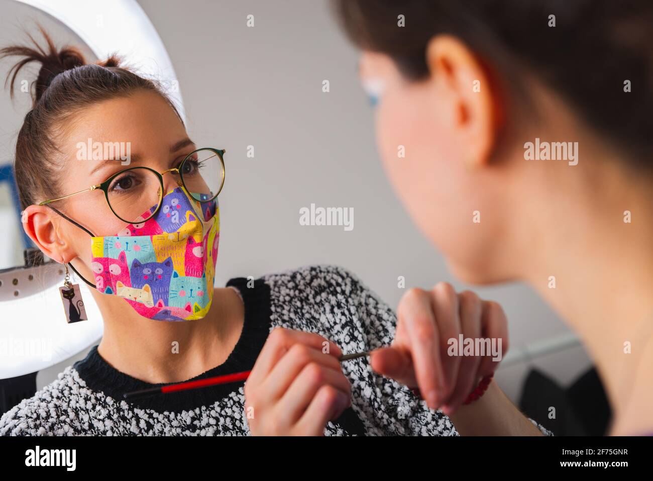 Primo piano di un estetista che indossa una colorata maschera protettiva durante l'applicazione make up su bella giovane donna in sedia Foto Stock