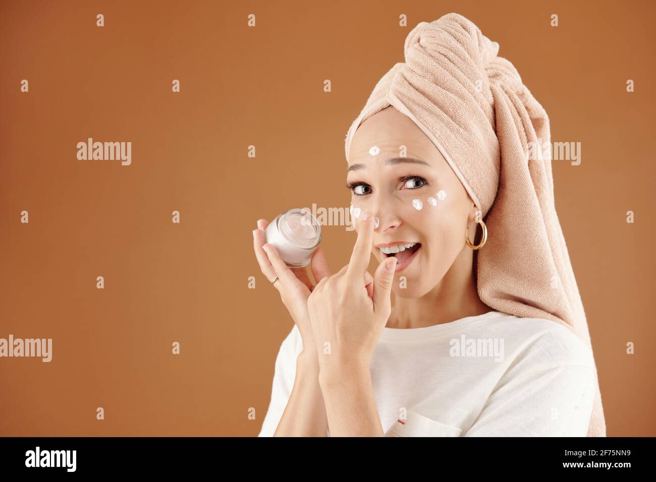Felice giovane donna eccitata con asciugamano sulla sua testa che si applica lozione idratante dopo la presentazione mattutina Foto Stock