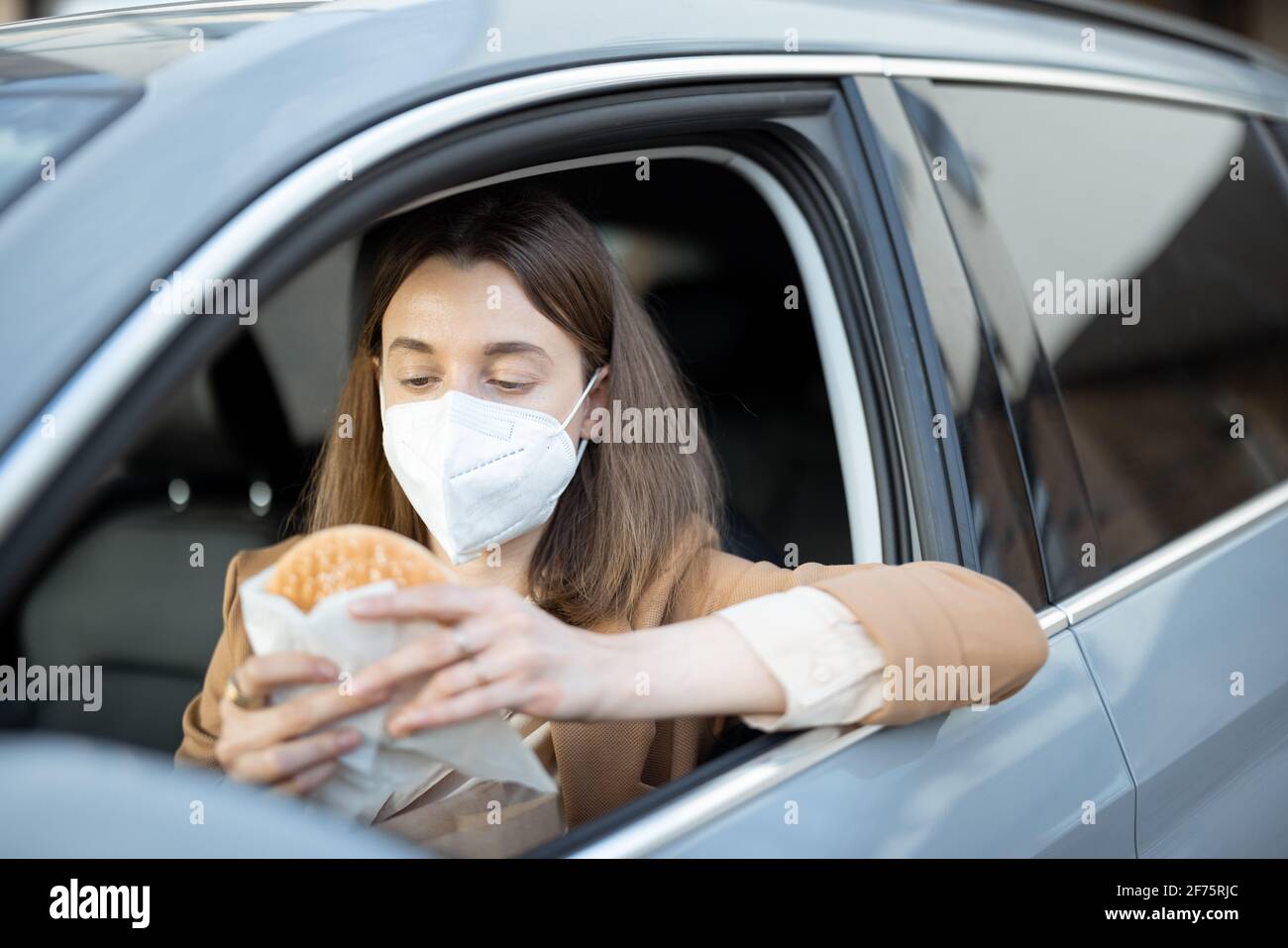 Giovane donna con maschera protettiva sul viso ha preso l'ordine del cibo per andare e ha ottenuto un hamburger dal pacchetto. Sanità, protezione da virus. Foto Stock