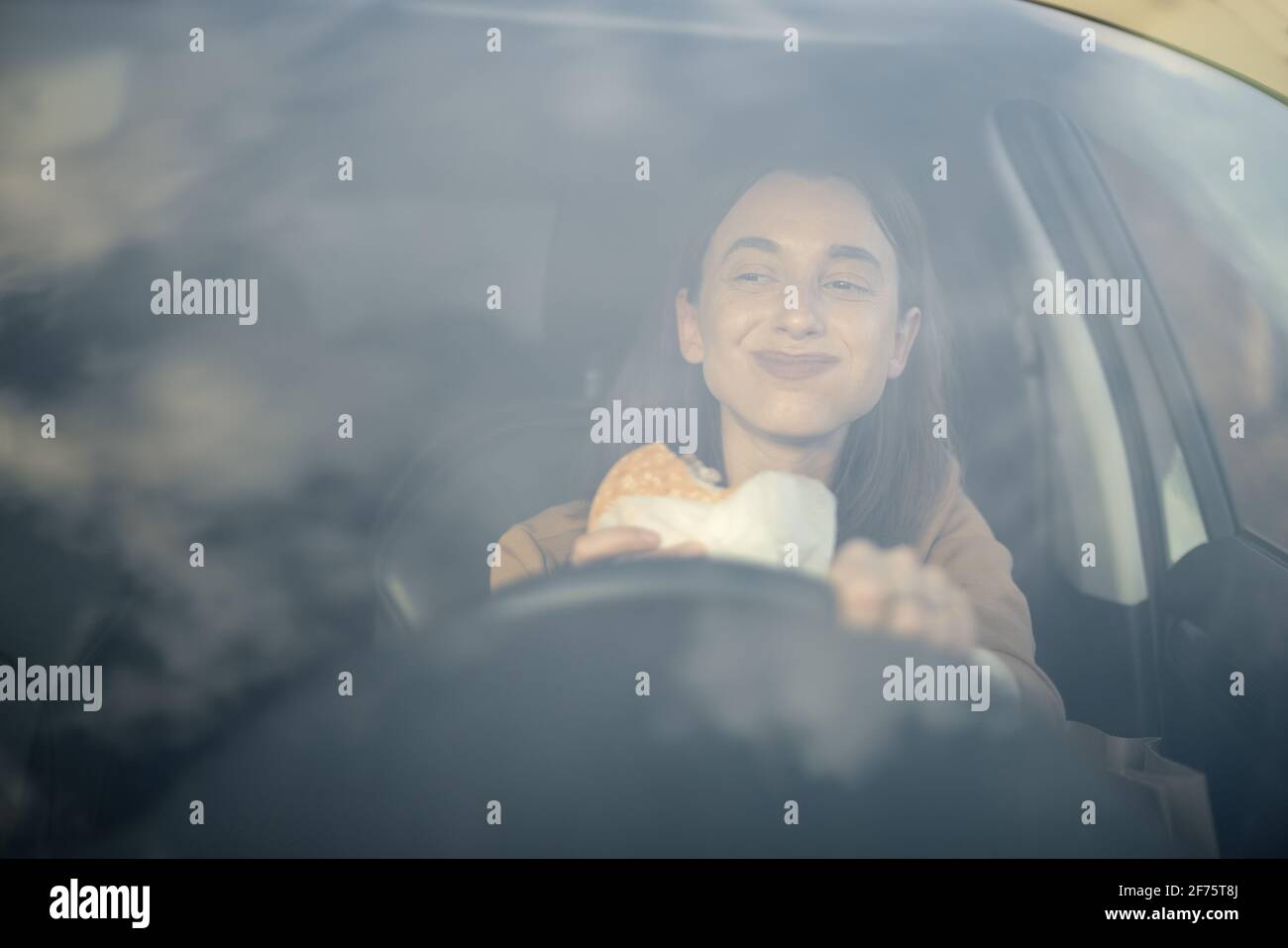 Donna felice che mangia un hamburger in auto mentre si guida e si guarda la strada. Avere uno spuntino fast food malsano. Cibo da andare. Concetto affamato e affollato. Foto Stock