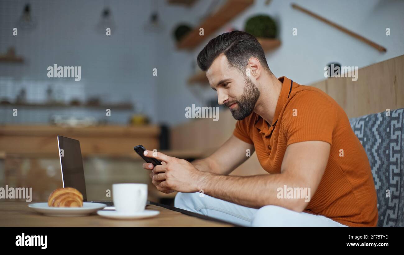 messaggi sorridenti di freelance sullo smartphone vicino al computer portatile e colazione in caffè Foto Stock