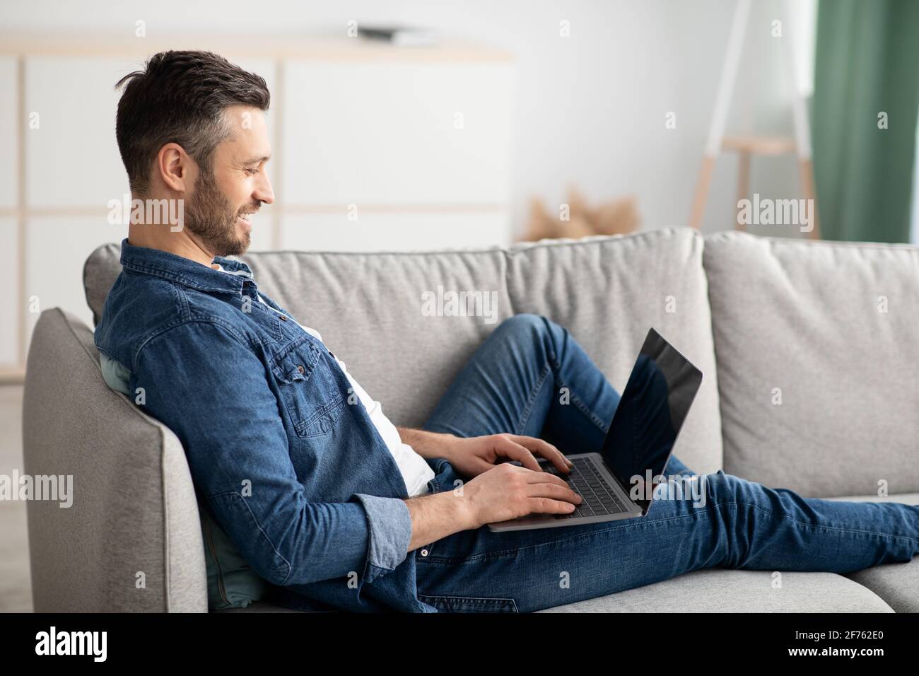 Vista laterale di un uomo con la bearded reclinato sul divano, utilizzando un computer portatile Foto Stock