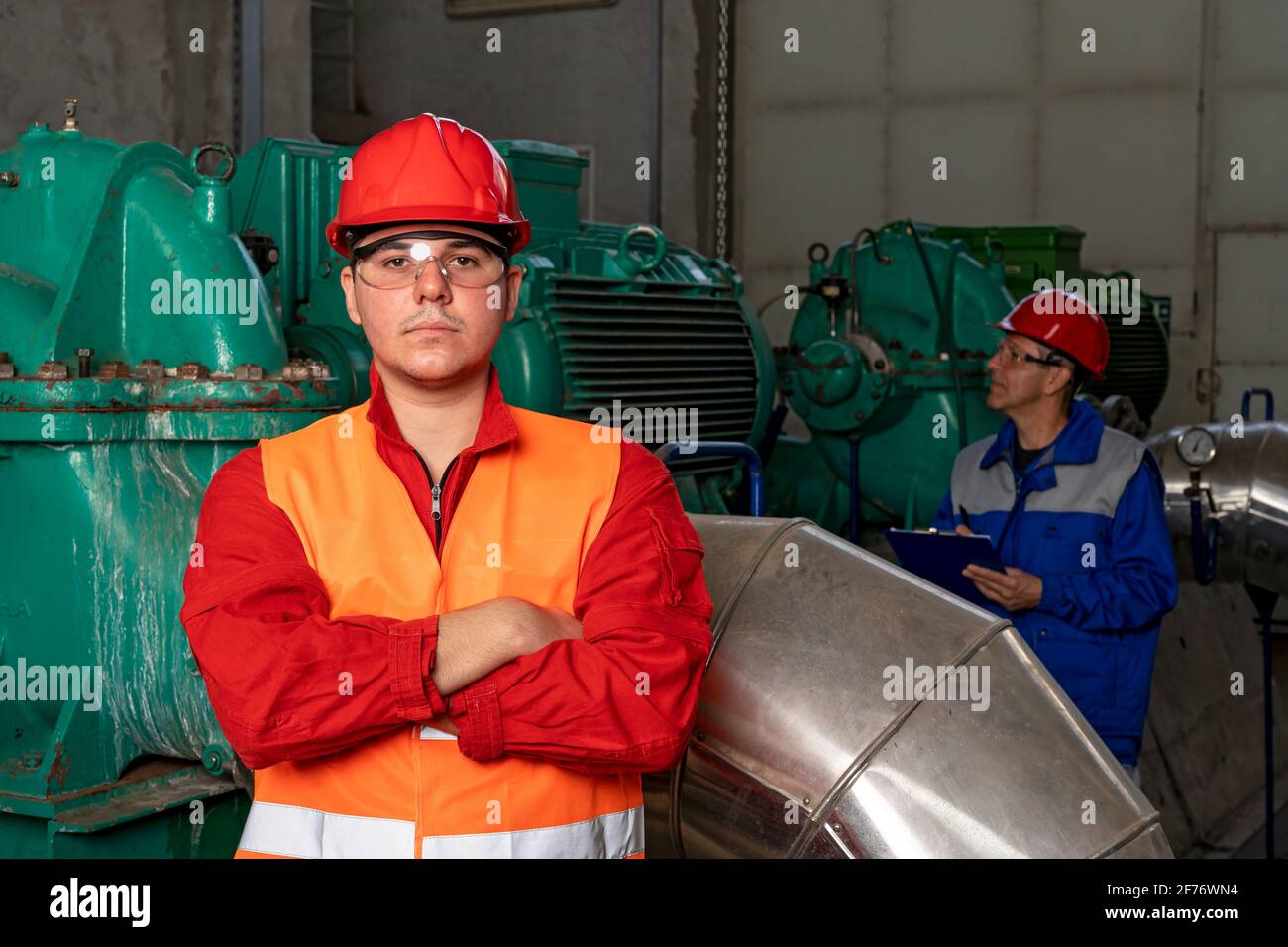 Giovane lavoratore in tute rosse e Hardhat in piedi in interni industriali con tubi e generatori. Ritratto di un operatore giovane sicuro. Foto Stock