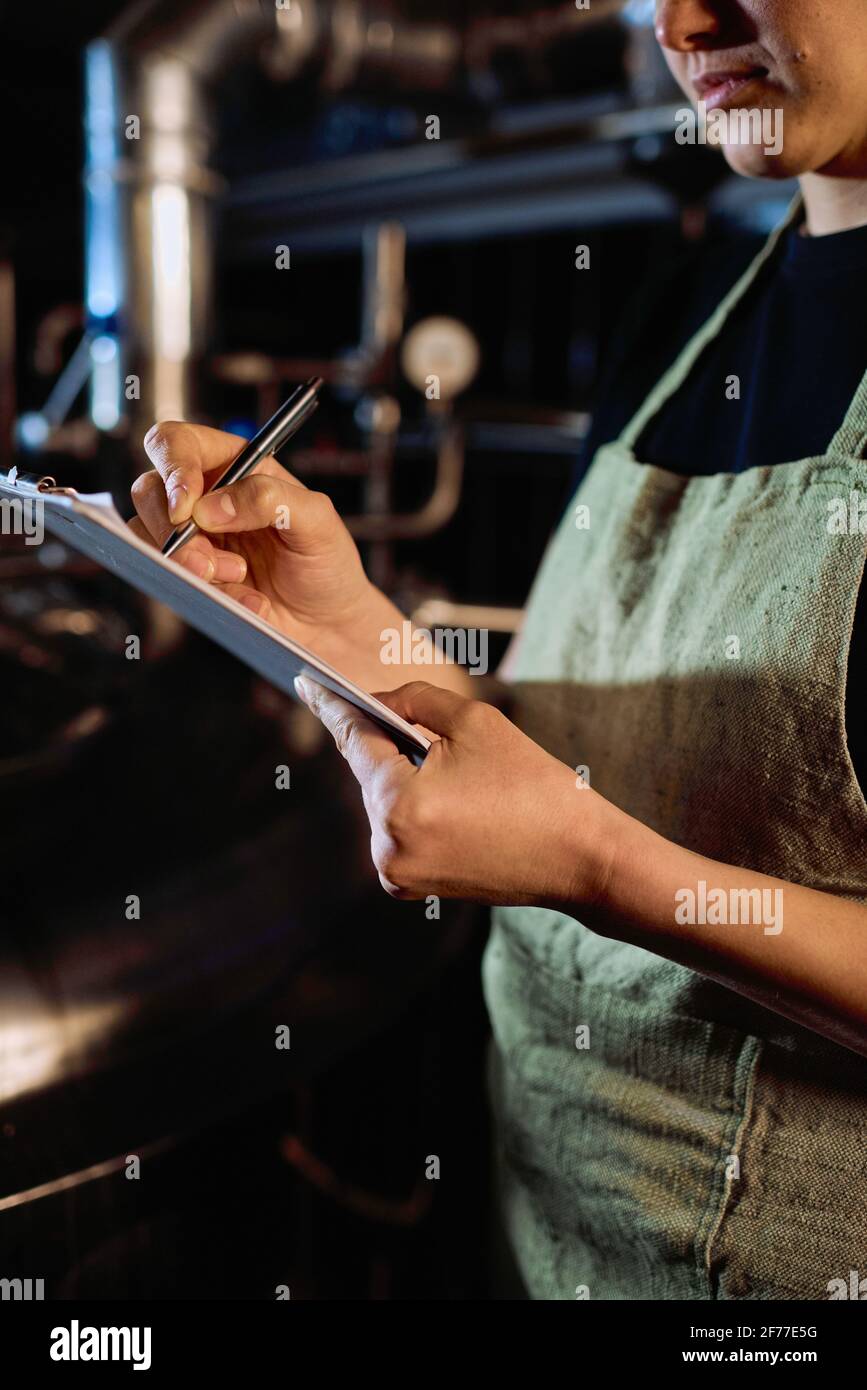 Giovane birraio in grembiule che punta al documento negli appunti durante l'apprendimento dell'elenco delle fasi di preparazione della birra alla produzione pianta Foto Stock