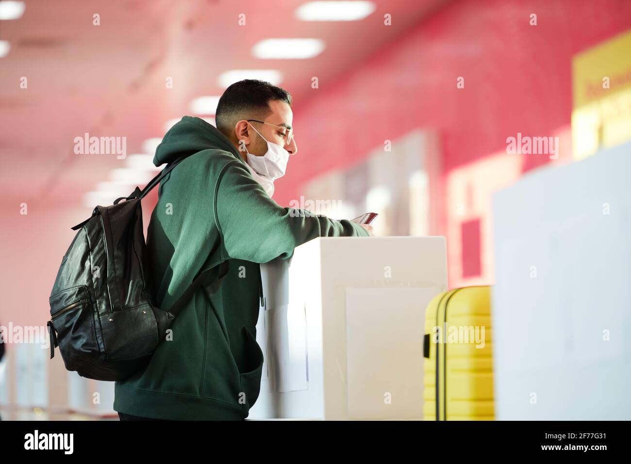 Giovane uomo in casualwear, occhiali e maschera protettiva in piedi presso il banco di registrazione nella sala dell'aeroporto e in attesa di receptionist Foto Stock