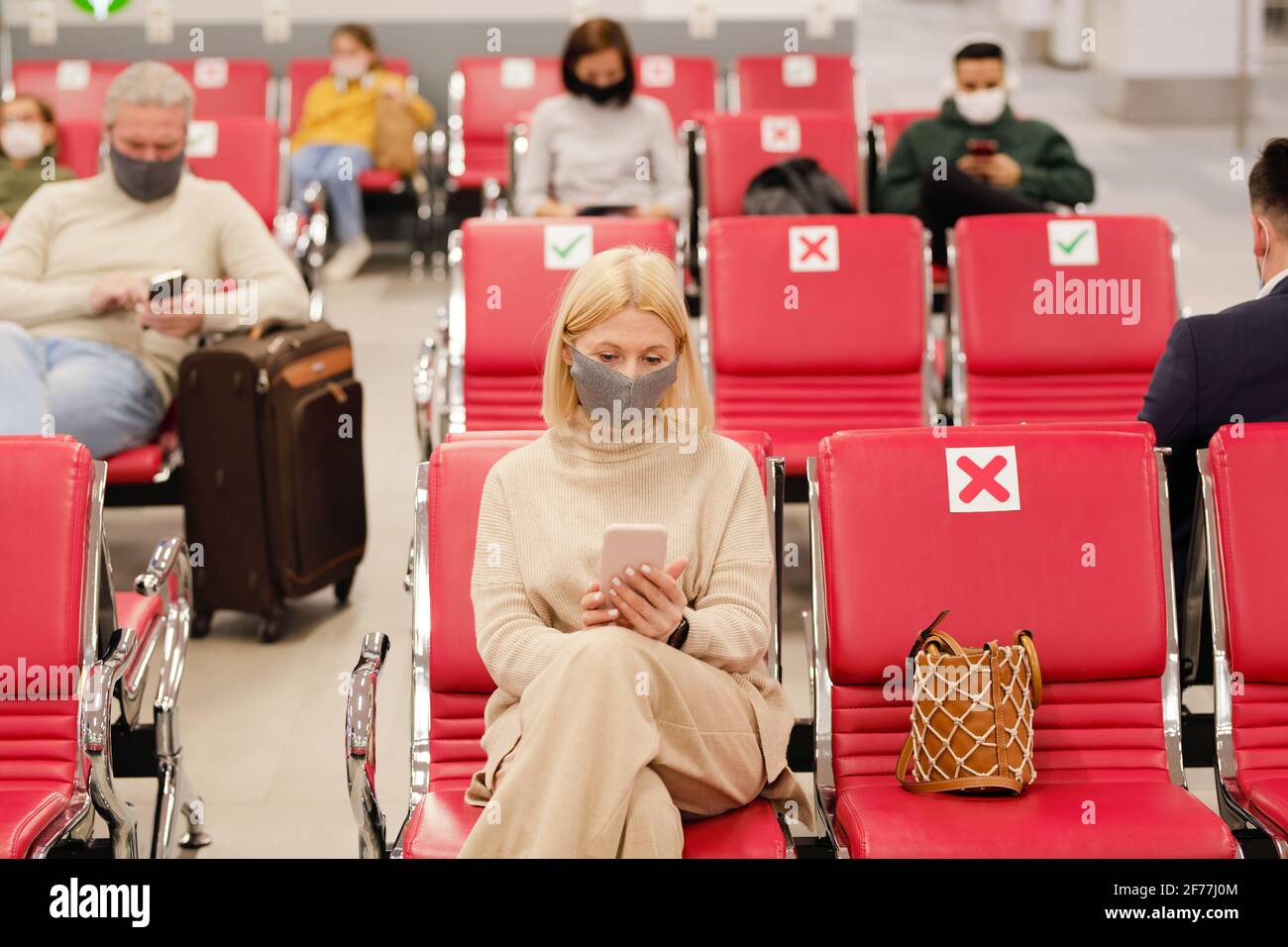 Ragazza bionda matura in casualwear e maschera protettiva utilizzando smartphone contro altre persone in attesa di annuncio del volo Foto Stock