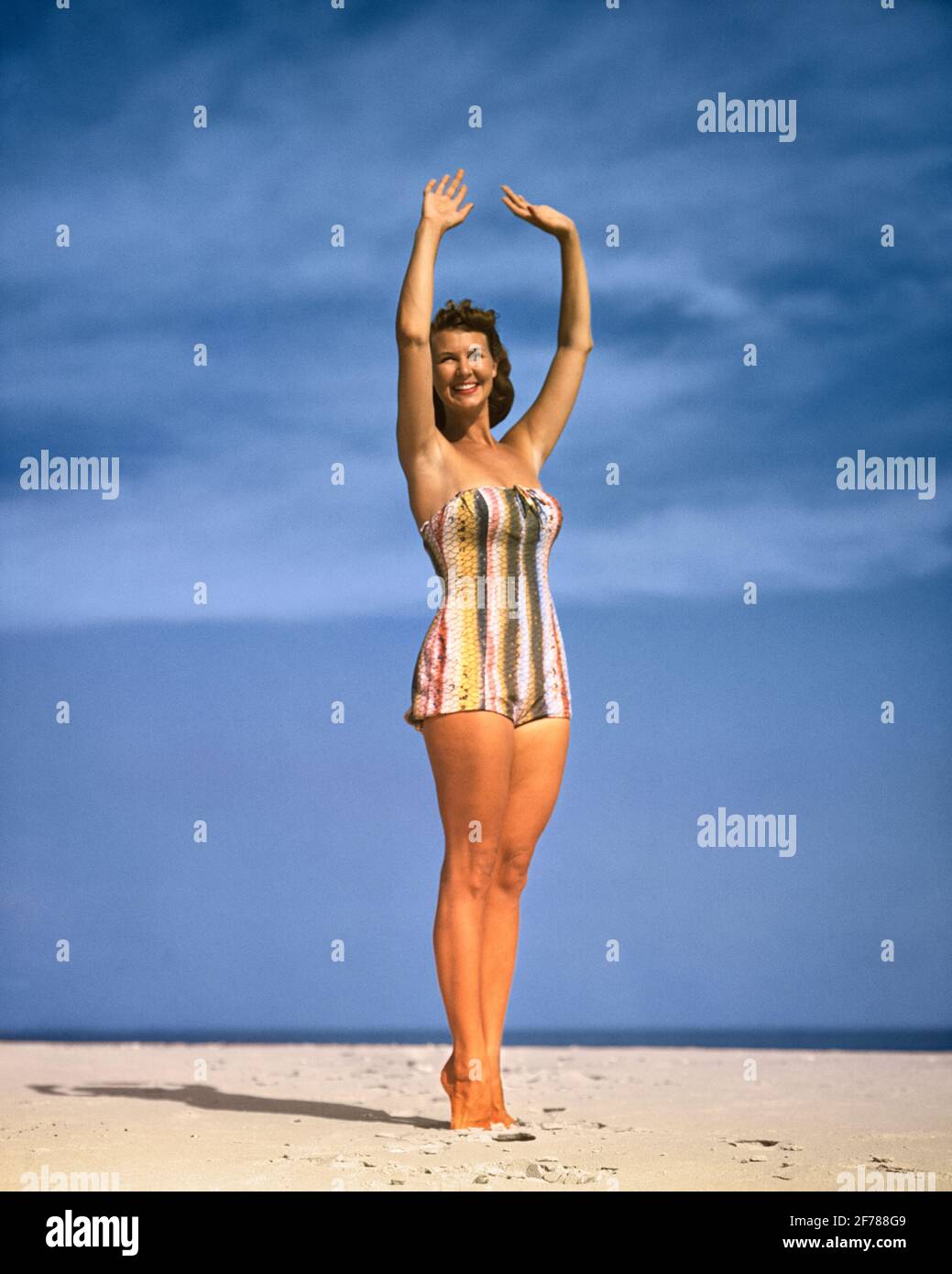 DONNA SORRIDENTE DEGLI ANNI '50 IN UN PEZZO DI BAGNO A RIGHE DI COLORE VERTICALE ABITO POSATO SU BEACH ARMS RAISED FT LAUDERDALE FLORIDA USA - KB32336 HAR001 HARS COPY SPACE FULL-LENGTH SIGNORE PERSONE OCCHIO CONTATTO BRUNETTE TEMPO OFF SHORE ALLEGRO STILI VIAGGIO FUGA RICREAZIONE SPIAGGE VACANZE OCCUPAZIONI POSANDO SORRISI GIOIOSO SABBIOSO ELEGANTE MODA MODELLARE POSA ABBRONZATO ABBRONZATO VACANZE GIOVANE DONNA ADULTA ETNIA CAUCASICA HAR001 VECCHIO STILE Foto Stock