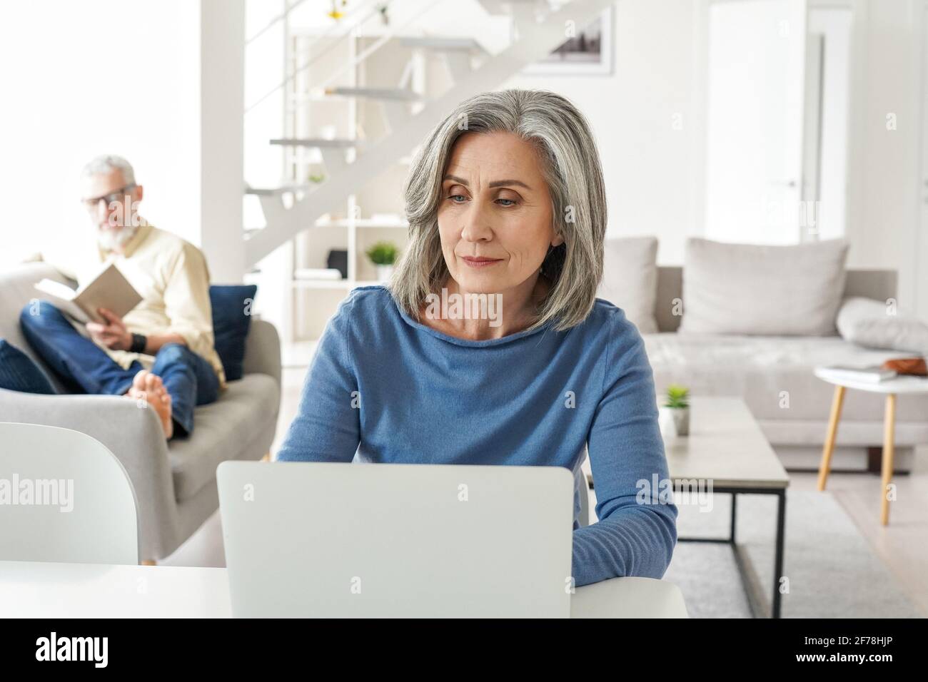Donna matura che lavora online da casa mentre il vecchio marito si rilassa in soggiorno. Foto Stock