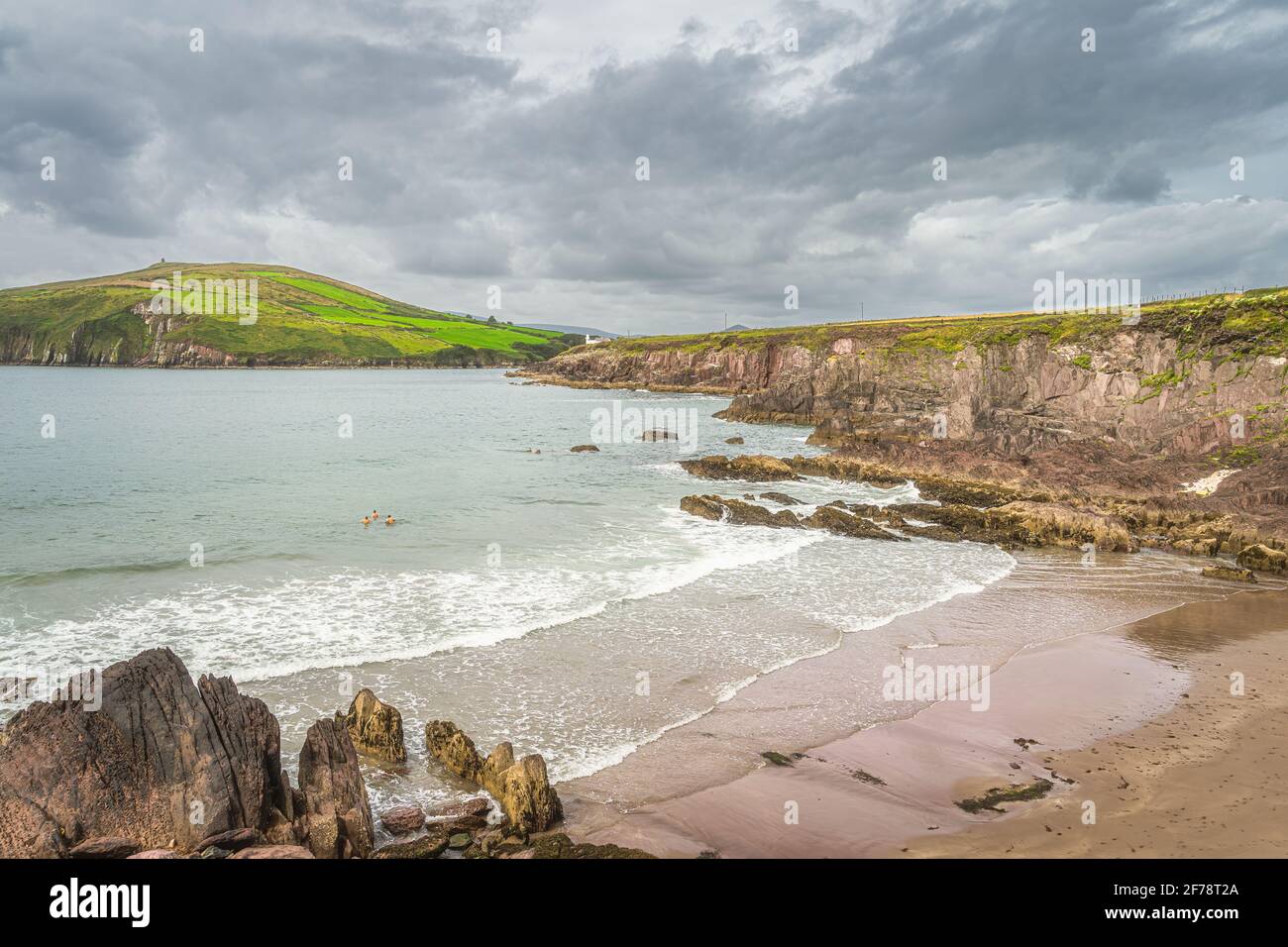 Persone che nuotano in acque fredde dell'Oceano Atlantico. Faro di Dingle sul bordo della scogliera con nubi tempesta sullo sfondo, Kerry, Irlanda Foto Stock