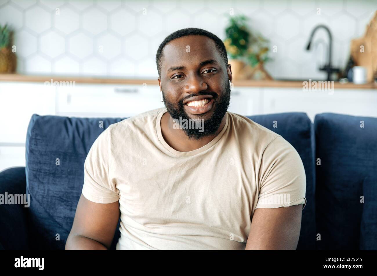 Primo piano ritratto di bel giovane sicuro afro-americano uomo bearned, seduto sul divano a casa in soggiorno, indossando t-shirt, guardando la macchina fotografica e sorridendo amichevole Foto Stock