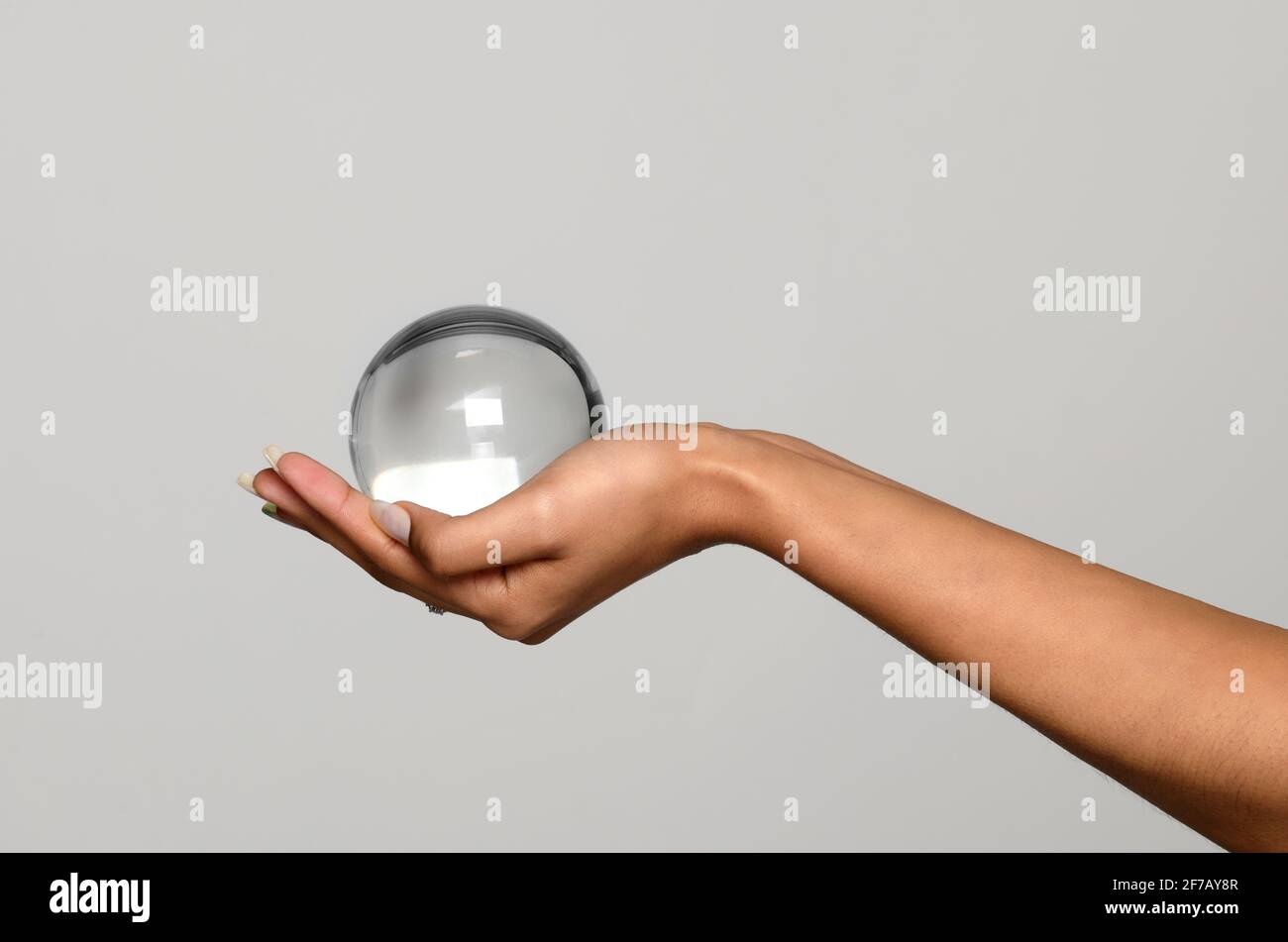 Mano di una giovane donna nera che tiene una sfera trasparente di vetro o cristallo, orb o palla imbutita nel suo palmo su uno sfondo grigio chiaro studio con poliziotto Foto Stock