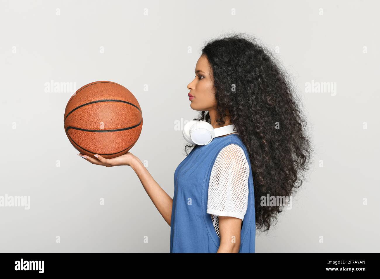 Profilo di una giovane donna nera in leisurewear con lunghi capelli ricci e cuffie intorno al collo che tengono un basket in mostra sul palmo di Foto Stock