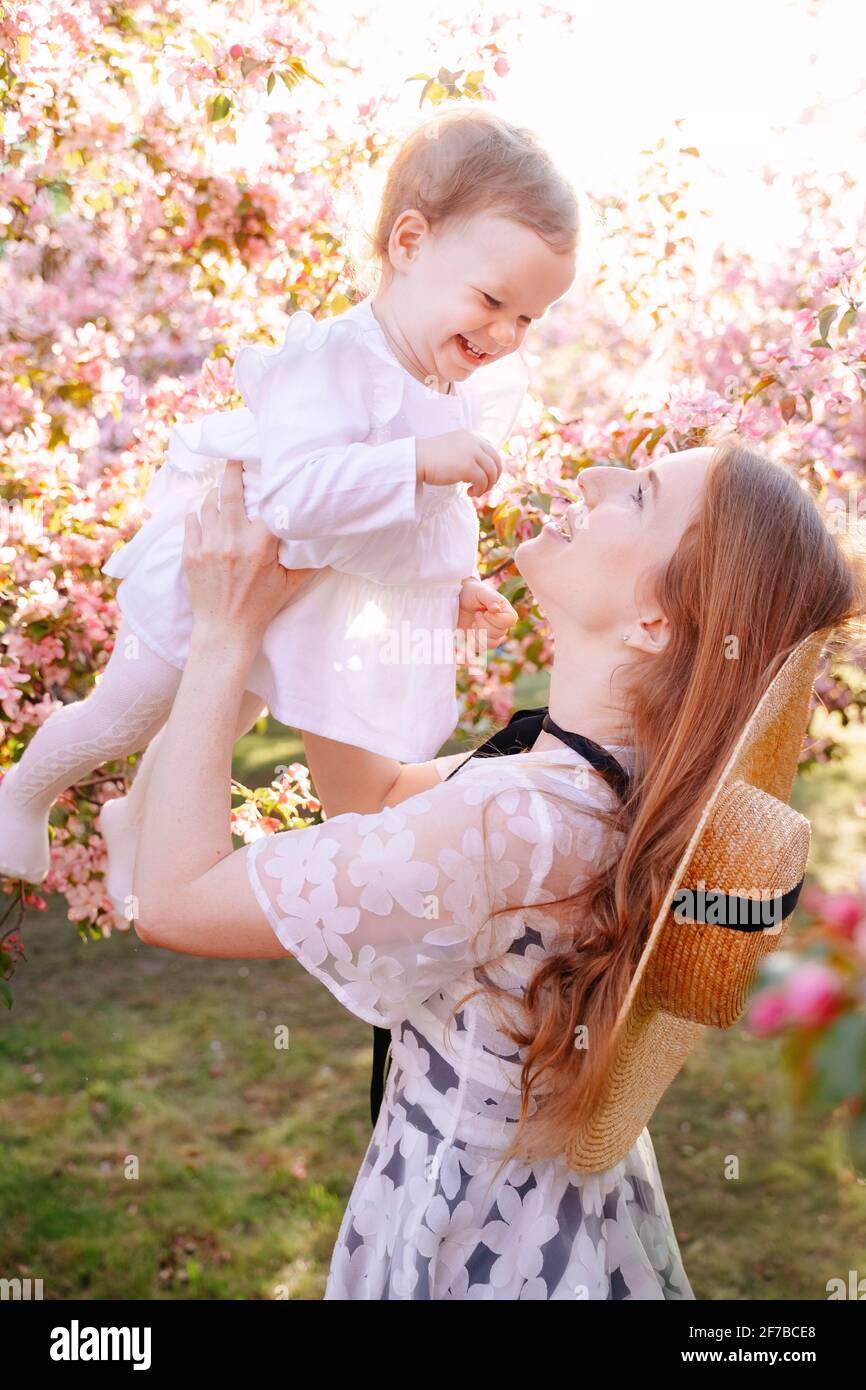 Un genitore tiene una bambina sorridente tra le braccia contro uno sfondo di un albero di rose in fiore. Foto Stock
