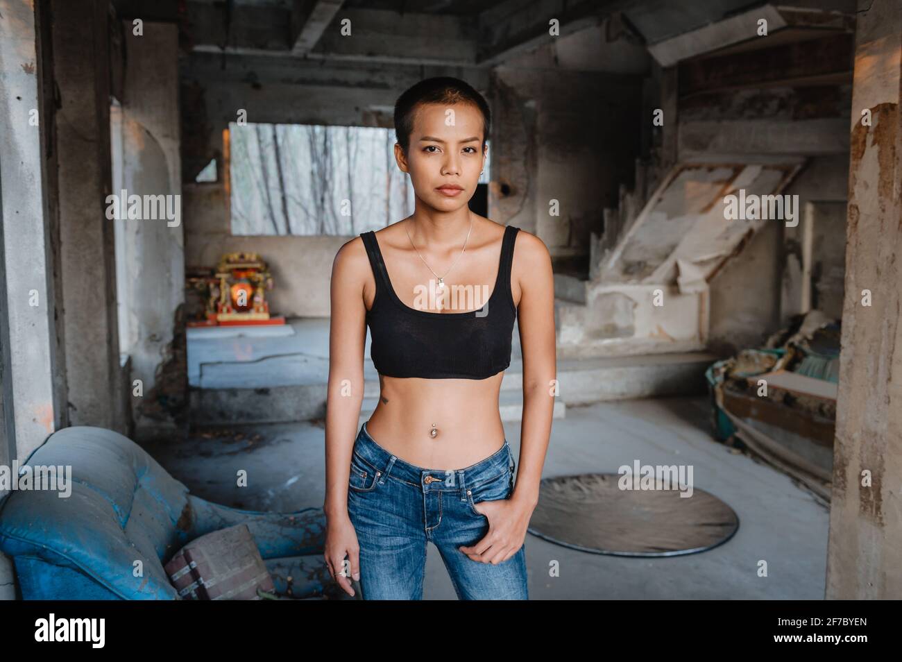 Una giovane donna asiatica capelli corti che indossa un reggiseno sportivo e jeans blu è in piedi in una casa abbandonata guardando la fotocamera. Foto Stock
