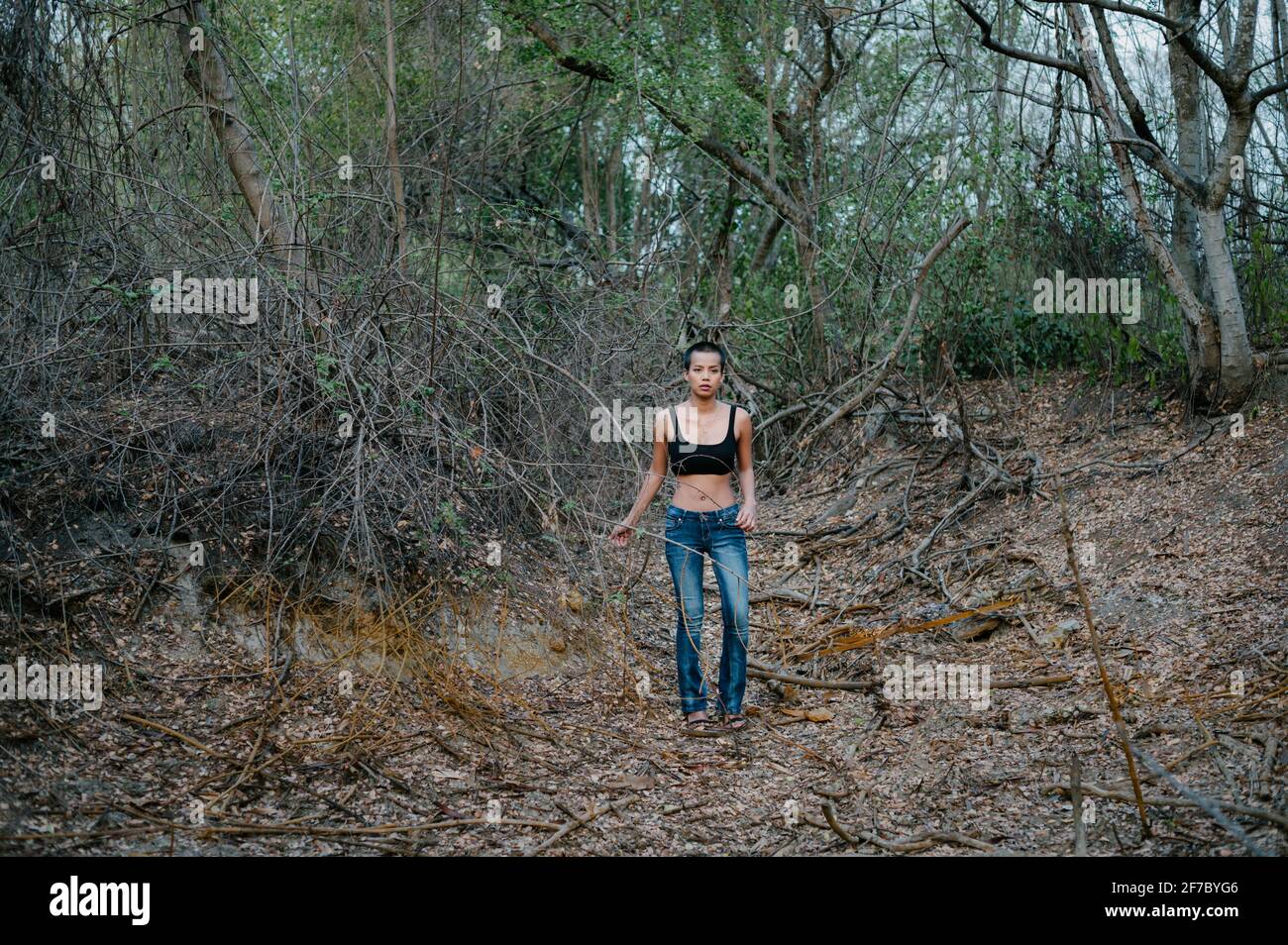Foto lunga di una giovane donna asiatica capelli corti in piedi da sola in una foresta, indossando un reggiseno sportivo e jeans blu Foto Stock