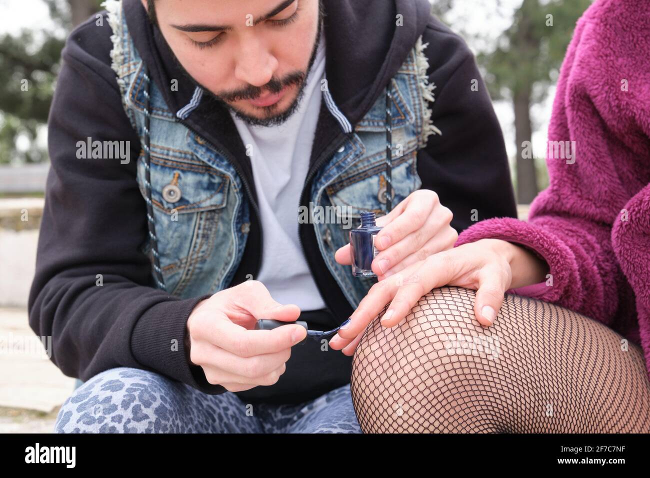 Giovane punk uomo dipingendo unghie alla sua amica in un parco. Due amici che lucidano le unghie all'esterno. Foto Stock