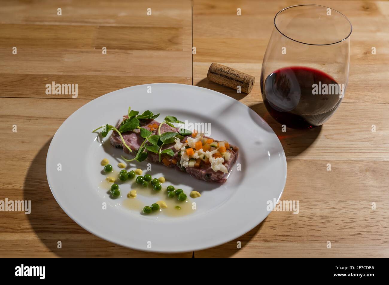 Menu degustazione raffinata portata da pranzo servita su piatto bianco: Terracina di prosciutto e porro con germogli di piselli, pease e sottaceti con bicchiere di vino rosso e sughero Foto Stock