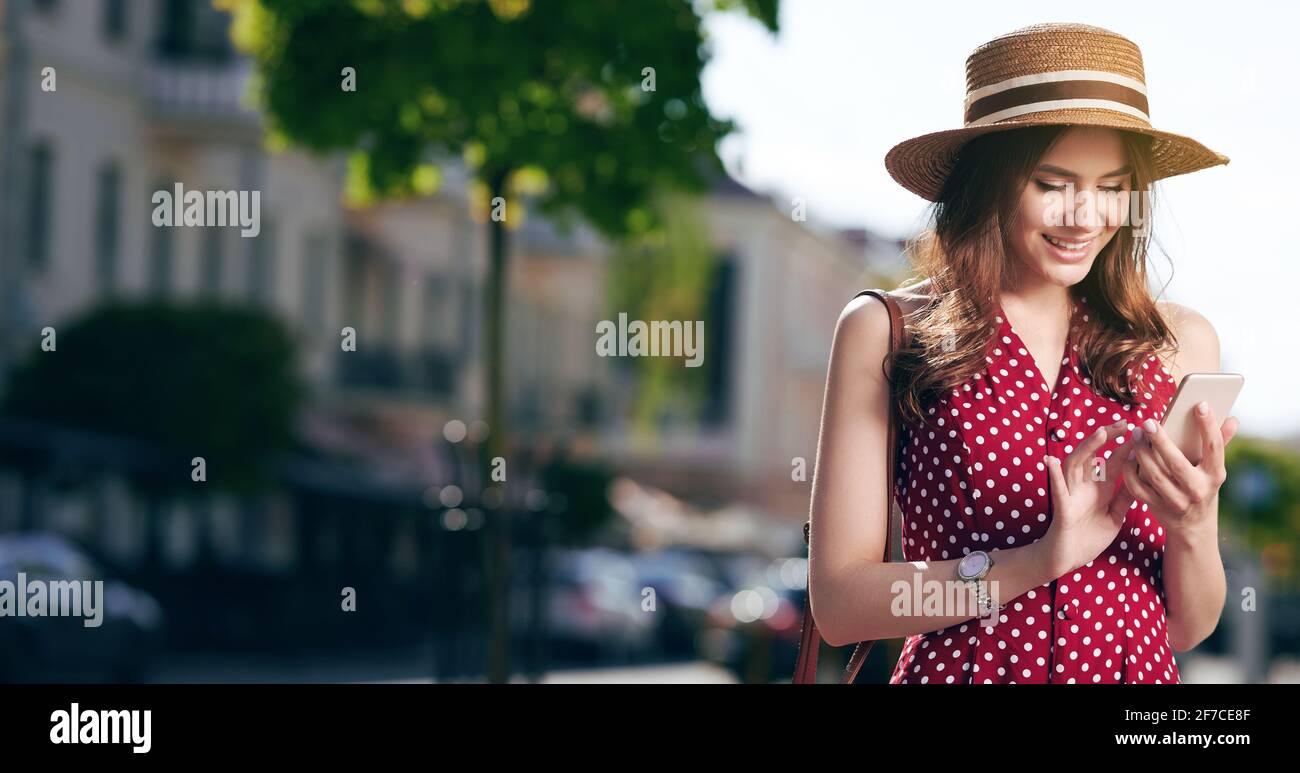 Caucasica donna scrive messaggi sul suo smartphone e entra la città estiva con un cappello di paglia e una bella abito rosso con pois di polka Foto Stock